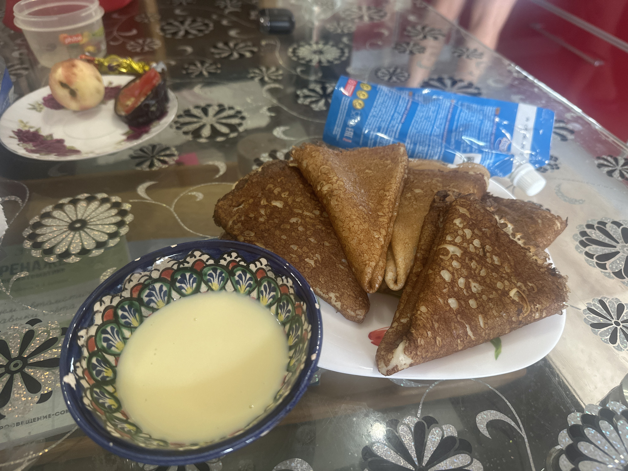 A close-up, eye-level shot captures a breakfast scene on a glass table in an indoor setting, possibly a dining area in Protvino, Russia. The table is adorned with a patterned tablecloth featuring floral motifs.
In the foreground, a small, blue and white decorative bowl is filled with a pale yellow, creamy liquid, likely condensed milk or a similar sweet sauce. To its right, a white plate holds several folded crepes, browned to a golden hue, some with visible fillings. One crepe is partially unfolded, revealing a light-colored interior.
Behind the crepes, a blue and white plastic pouch, possibly containing jam or syrup, lies on its side with its cap slightly ajar. A small white saucer sits to the left of the crepes and slightly behind the bowl. On this saucer are two pieces of fruit: one appears to be a whole peach, and the other is a slice of dark fruit, possibly a plum or fig, with a red center.
In the upper left corner, a clear plastic container with a white lid is visible, containing a light-colored liquid. The background is blurred, but hints of red furniture and legs of a person are discernible, suggesting the presence of other individuals in the room. The overall impression is one of a casual, home-cooked meal.