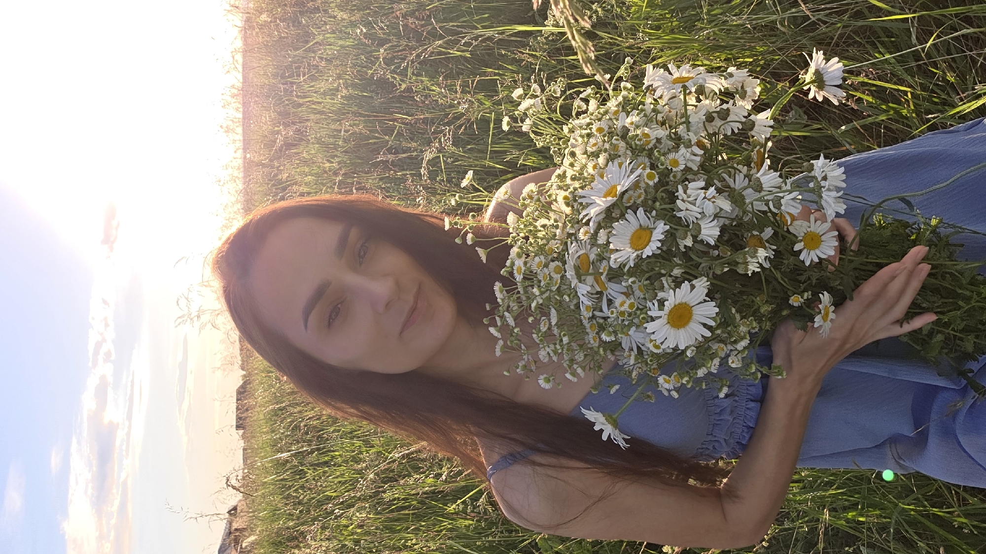 The image depicts a young woman holding a large bouquet of daisies in a field of tall grass, likely during sunset. The sun is low on the horizon, casting a warm glow and creating lens flare in the upper left portion of the image. The woman has long, dark hair and is wearing a blue dress with thin straps. Her face is partially obscured by the flowers, but she appears to be looking towards the camera with a gentle expression. The scene is set in a natural outdoor environment, with the tall grass and wildflowers suggesting a rural or countryside location, possibly in Купоярово, Russia, given the context. The overall mood is serene and peaceful, evoked by the soft lighting, the natural setting, and the woman's relaxed posture. No text is visible in the image.