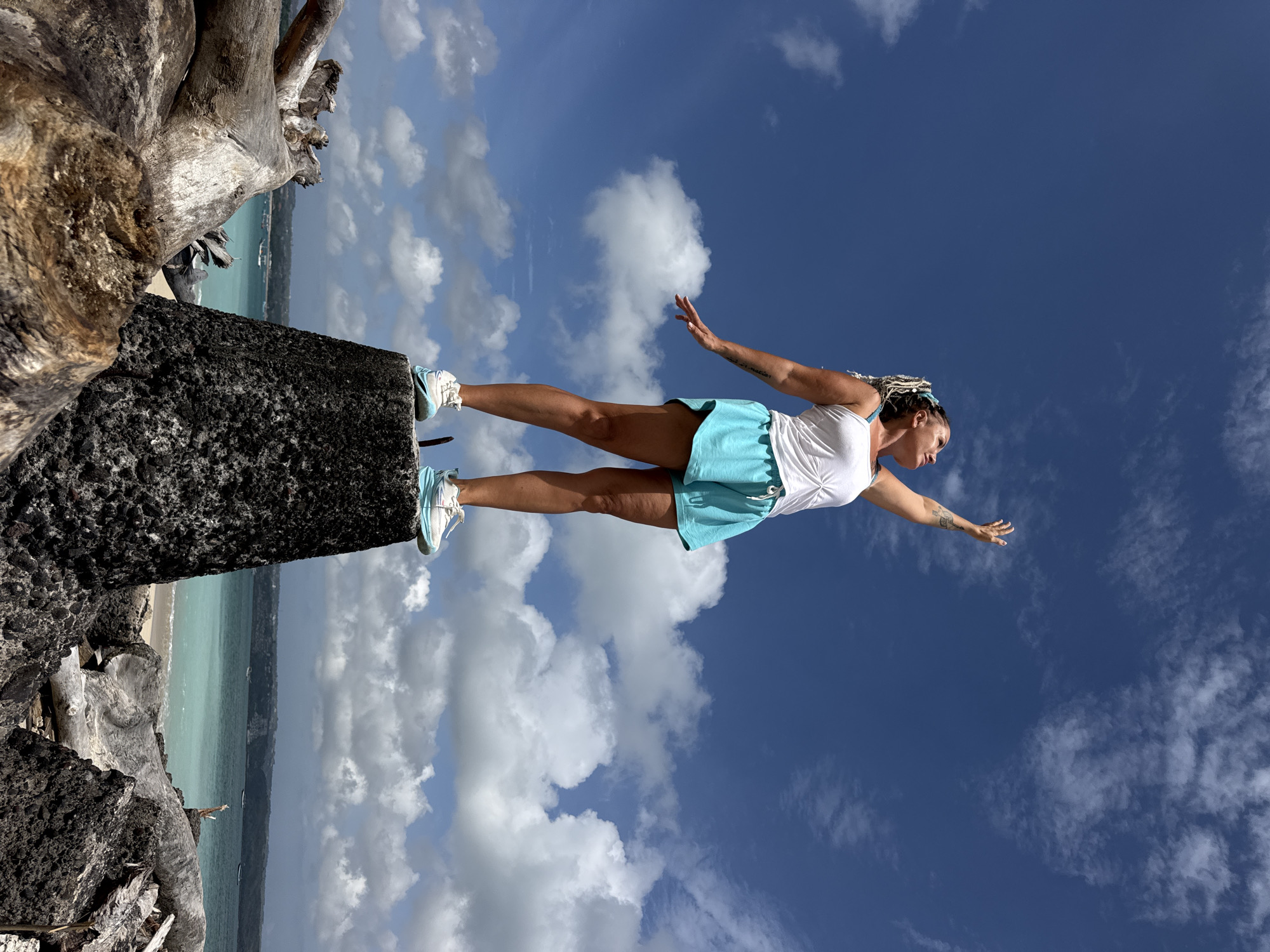 A woman stands with her arms outstretched on top of a dark, conical structure. She is wearing a white tank top, teal shorts, and athletic shoes. Her hair is styled in blonde dreadlocks. The structure she stands on is rough and dark, possibly volcanic rock or concrete. In the foreground, large pieces of weathered driftwood are scattered. Behind her, a bright turquoise ocean meets a sandy beach, with a distant coastline and a few boats visible. The sky is a vivid blue with scattered white clouds. The scene appears to be a coastal area, possibly near Denpasar, Indonesia, given the typical geography and vibrant ocean colors.