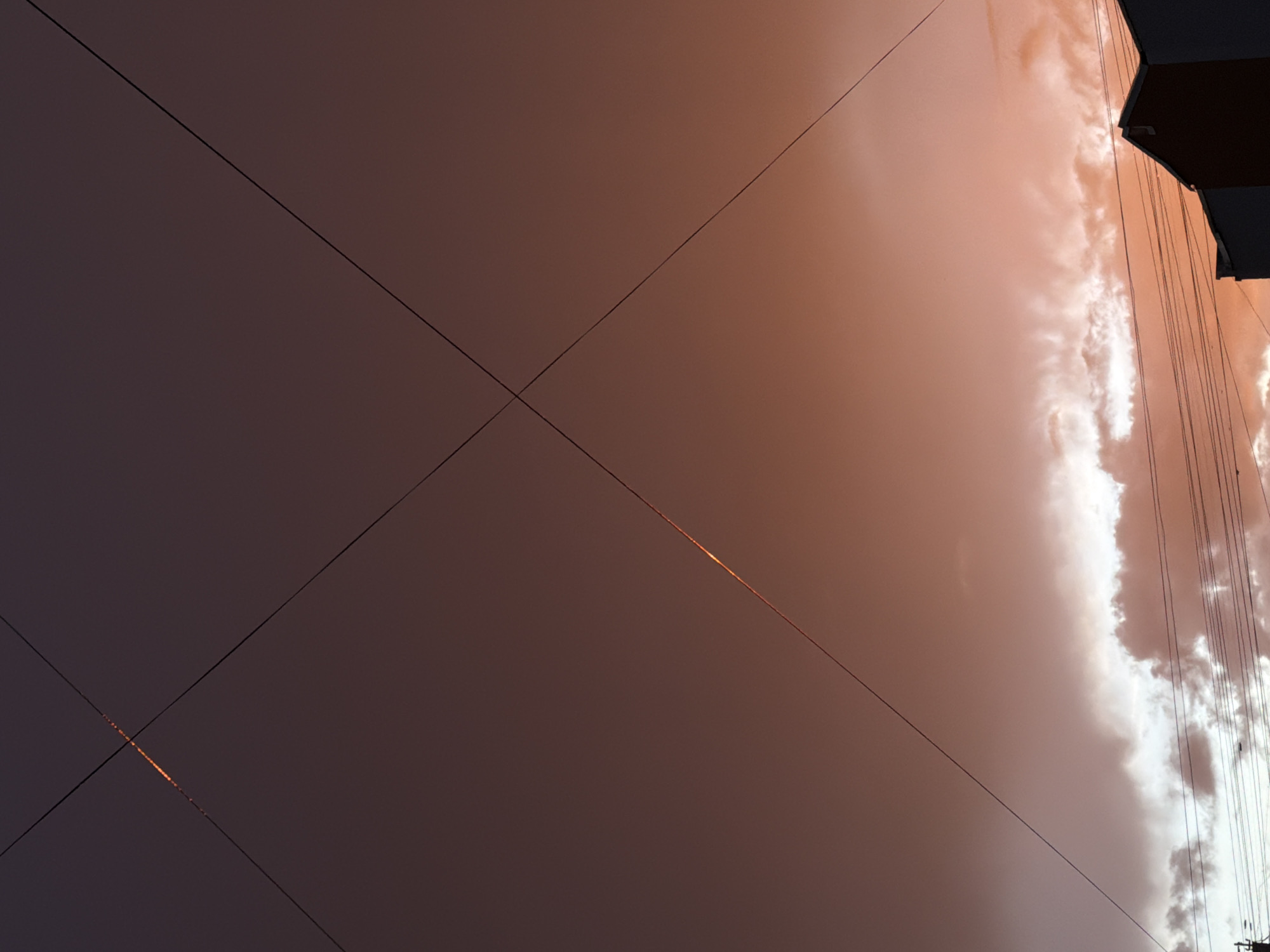 The image shows a dusky sky over Khimki, Russia, with a dramatic sunset casting warm hues of orange and pink across the clouds. Two dark power lines bisect the sky diagonally, creating an X-shape formation. A thin, bright line of light is visible along the upper intersection of these power lines, possibly indicating sunlight catching the wires or a distant light source. Below the sky, the silhouette of a building's rooftop is visible on the right side, with more power lines running horizontally across the frame. The overall scene is one of urban twilight, with the celestial display dominating the view.
