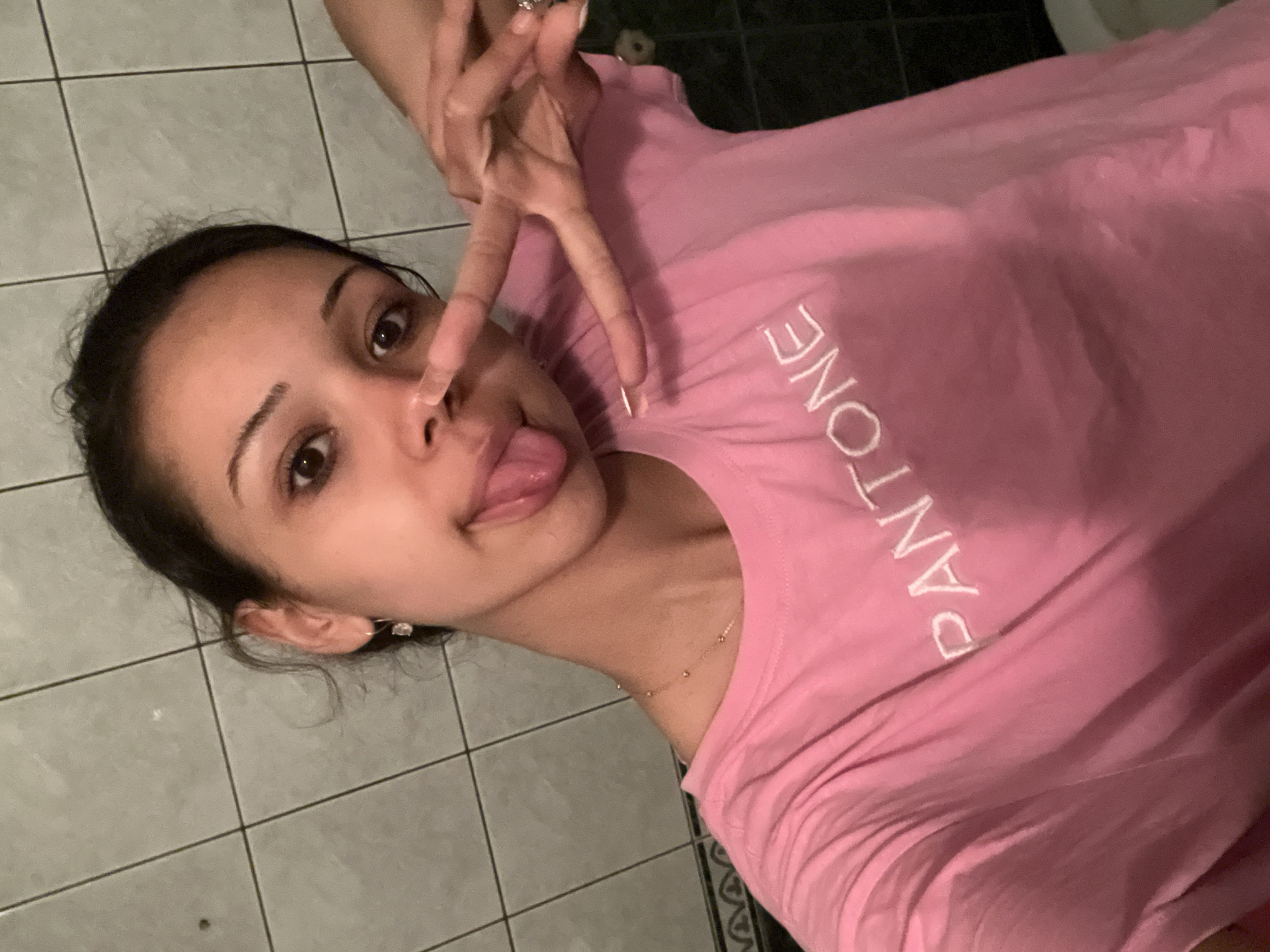 A young female, likely in her late teens or early twenties, with dark hair pulled back, light brown eyes, and fair to medium skin, is centered in a selfie-style portrait. She is making a playful expression with her tongue slightly extended and forming a peace sign (V-sign) with her left hand, which is held close to her face. She wears a light pink t-shirt with the word "PANTONE" embroidered in white across the chest. A delicate gold-colored necklace with small bead-like accents is visible around her neck, and a small, clear gemstone earring hangs from her right earlobe. Her fingernails are painted with a light, possibly metallic, polish. The background consists of a light gray tiled wall with dark grout lines, suggesting an indoor location, possibly a bathroom, given a faint glimpse of what appears to be a white toilet fixture in the lower right corner. The lighting is artificial and appears to be coming from above or the front. The image captures a candid, informal moment in the city of Unknown, Unknown.