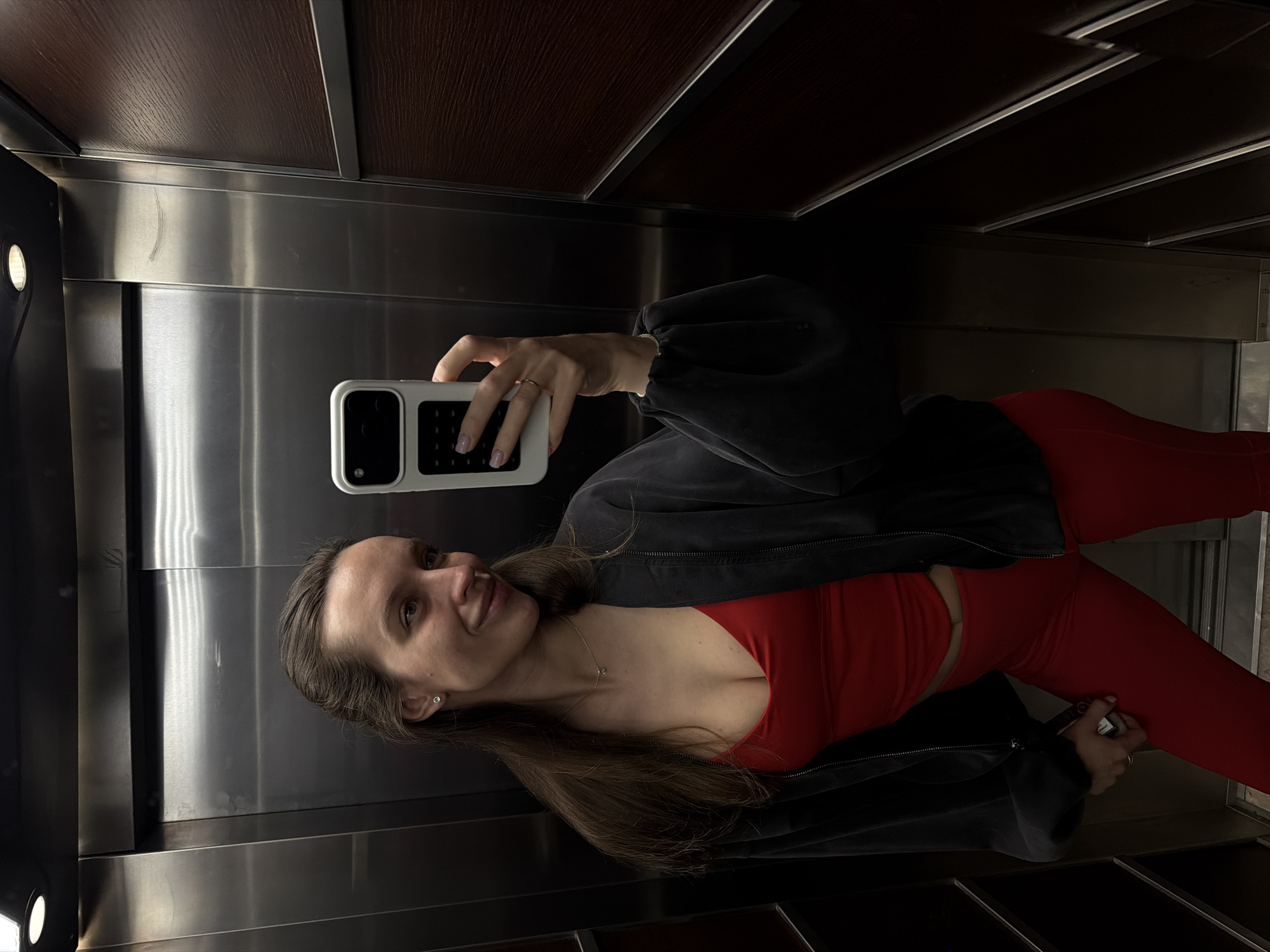 A woman is taking a selfie in an elevator. She is wearing a red top and leggings with a dark jacket. She is holding a phone with a white case. The elevator has metal walls and a wooden ceiling. It is unknown if the elevator is in Budva, Montenegro.