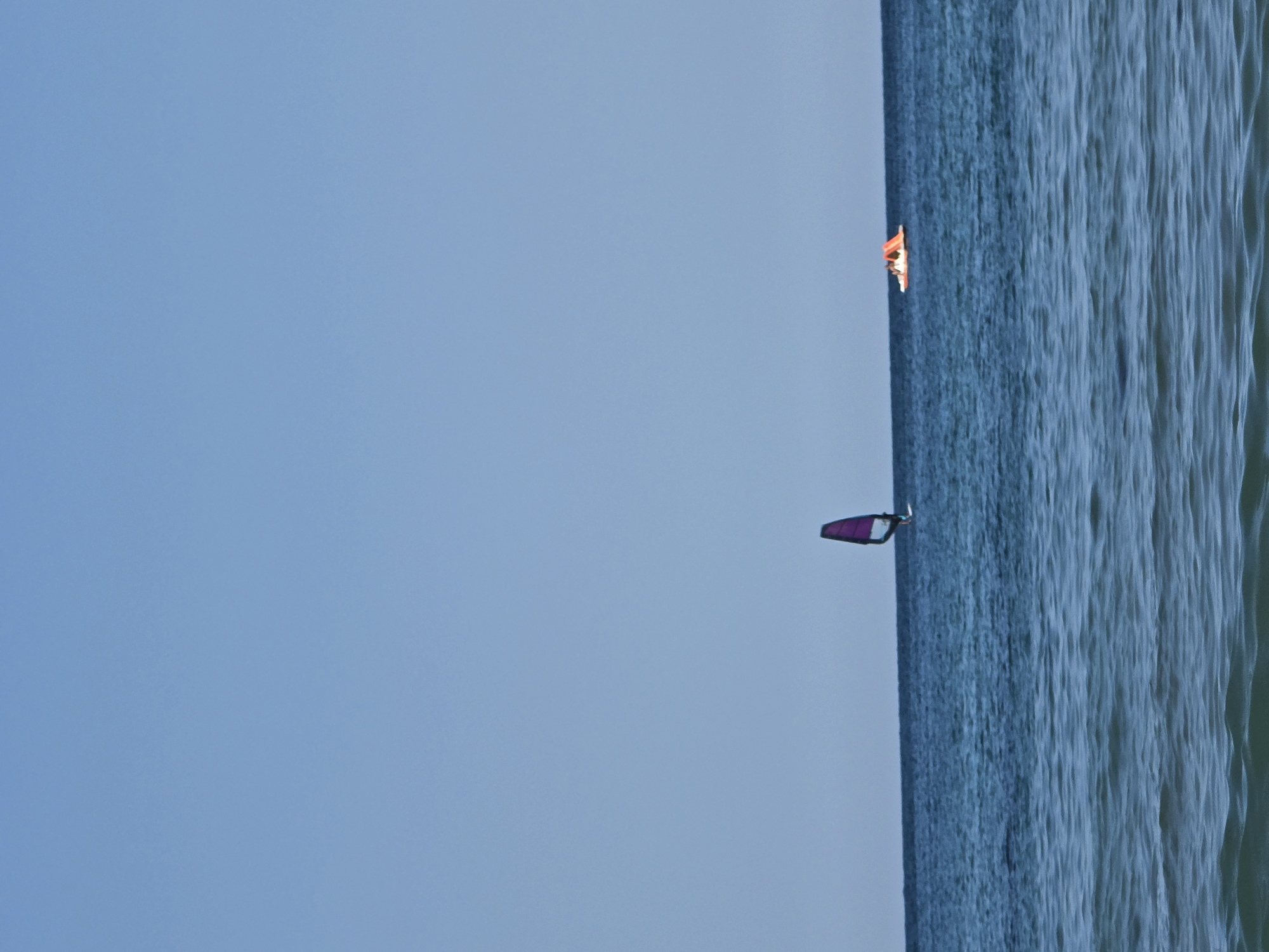 The image depicts a clear blue sky over a calm sea. In the mid-distance, a single windsurfer with a purple sail is visible on the water. Further to the right, a small orange and white pedal boat with at least two people on it is also present on the sea. The foreground shows the gentle ripples of the ocean. This scene is characteristic of the coastline near Ahtopol, Bulgaria, a popular destination for water sports and beach activities.