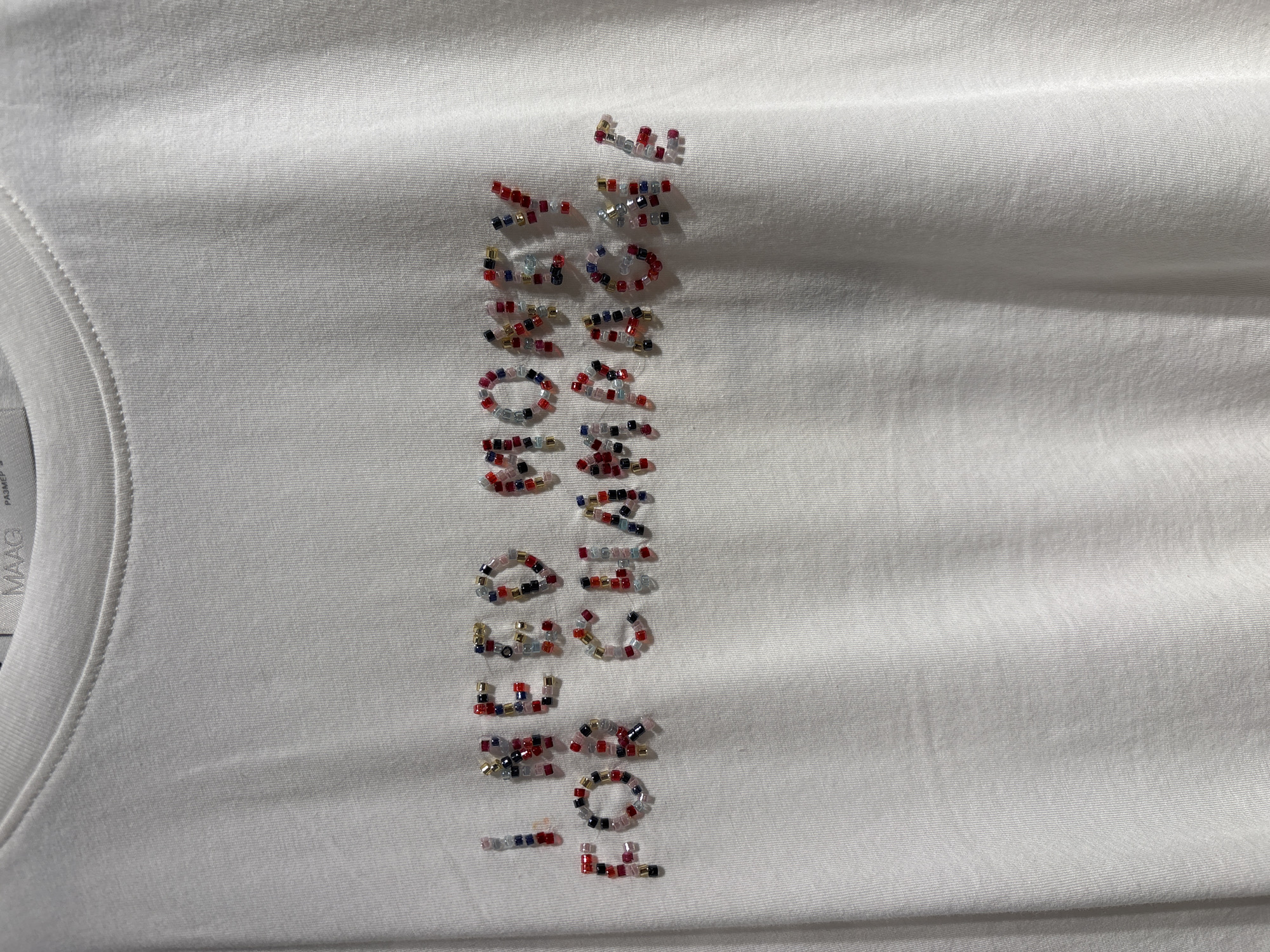 The image shows a white t-shirt with the phrase "You need somebody" spelled out vertically using colorful beads. The brand name "MAAG" is visible on a label near the collar. The location is not specified.