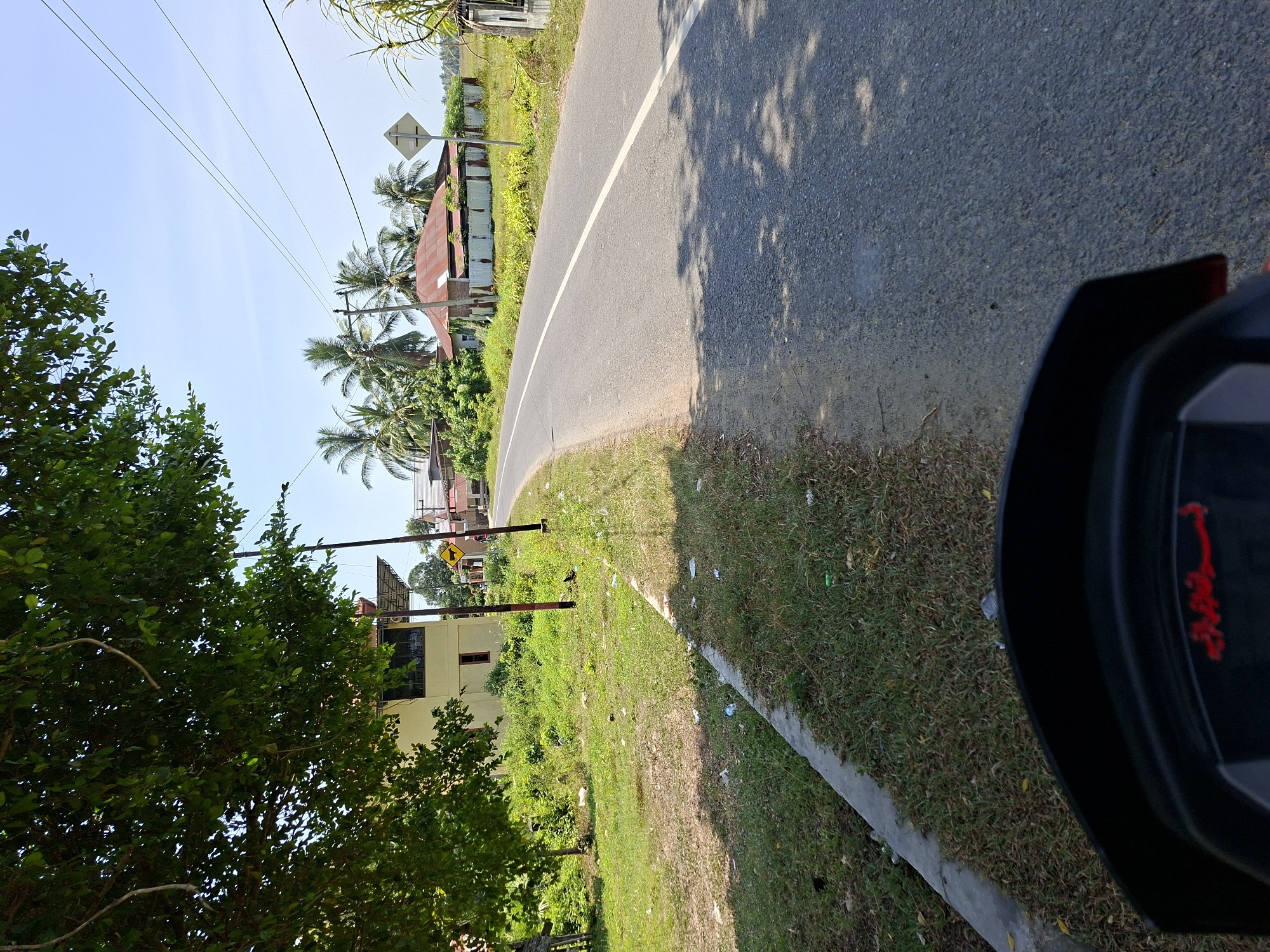 The image depicts a roadside scene in Mesjid, Indonesia, on a sunny day. The foreground shows the handlebars and dashboard of a motorcycle, with some red Arabic text visible on the dashboard. The road itself is paved and has a white dividing line. To the left of the road, there is a grassy area with some scattered litter and a concrete curb. A large tree with green leaves extends from the left side of the frame, casting shadows on the road.
Further down the road, on the right side, are several houses with corrugated metal roofs, surrounded by lush green vegetation and palm trees. A utility pole with electrical wires stretches across the scene. A diamond-shaped traffic sign with a black arrow indicating a curve is visible on the right side of the road. The sky is clear and blue. The overall impression is one of a typical rural or semi-urban road in Indonesia.