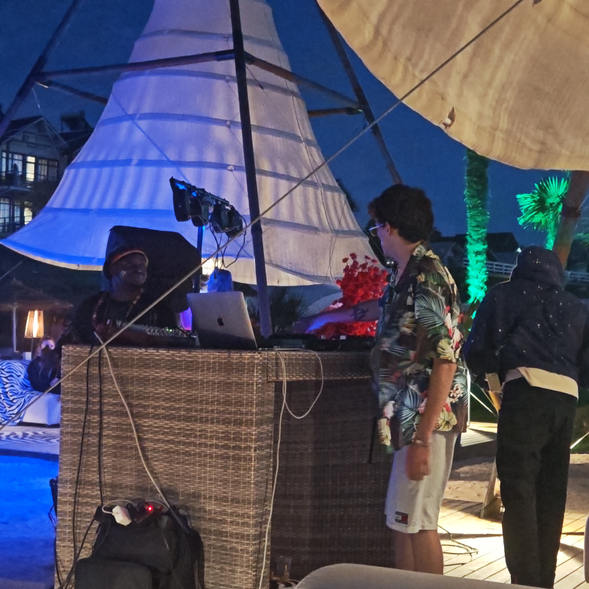 The image depicts an outdoor scene at night, possibly in a resort or beach area, with a DJ setup. Two individuals are visible, along with DJ equipment and ambient lighting.
The main subject on the left is a person wearing a hat and a dark t-shirt, appearing to be a DJ. They are positioned behind a woven wicker DJ booth, where a laptop and other equipment are set up. The DJ booth is illuminated by lighting from above and possibly from the equipment itself. There are cables and a power strip with a red indicator light visible on the DJ booth. To the left of the DJ booth, a lamp with a warm glow is present. A lounge chair with a zebra-patterned cushion is partially visible in the foreground to the left.
On the right, another person is seen from the back, wearing a short-sleeved, colorful tropical print shirt and light-colored shorts. This person appears to be interacting with the DJ or observing the setup. Further to the right, a third individual is partially visible, wearing a dark hooded jacket and dark pants. This person is also facing away from the camera.
The environment features a large, sail-like structure made of white fabric, illuminated with blue light from within. Another large, light-colored fabric structure, possibly an umbrella or canopy, is visible in the upper right corner. Palm trees are present, with one lit up in green light. Buildings, potentially part of a resort, are visible in the background. The ground appears to be made of wooden planks. The overall atmosphere suggests a nighttime entertainment or party setting. The location is identified as Мысовское сельское поселение, Ukraine.