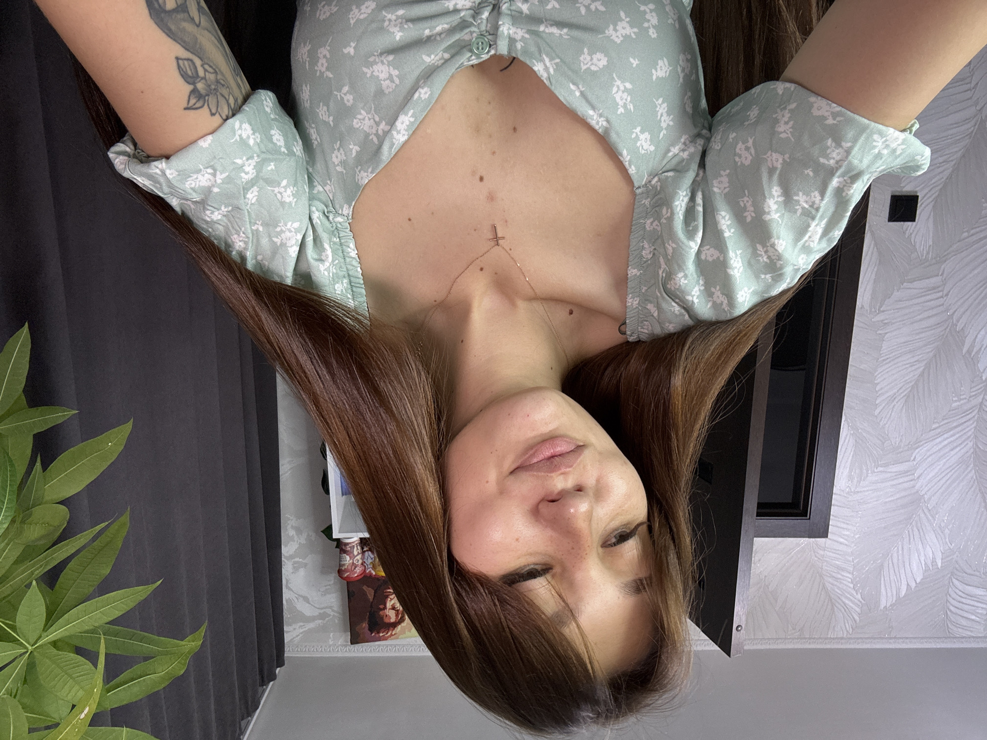 The image shows a close-up, head-and-shoulders portrait of a young woman with long, brown hair and fair skin, freckles, and dark eyebrows. She is wearing a light green, short-sleeved dress with a floral pattern and a small, gold cross necklace. Her hair is styled with bangs and falls loosely around her shoulders. She is looking down and to her left, with a slight smile.
The background suggests an indoor setting. To the left, there is a dark wooden door and a wall with a textured white wallpaper featuring a leaf pattern. A black light switch is visible on the wall. To the right, a dark gray curtain hangs, and a potted plant with green leaves is partially visible in the upper right corner. Above and behind the woman, there is a shelf with several items, including what appears to be a framed picture of a man and two cups or containers.
There is no information in the image to indicate the location is the city of Novokuznetsk, Russia.