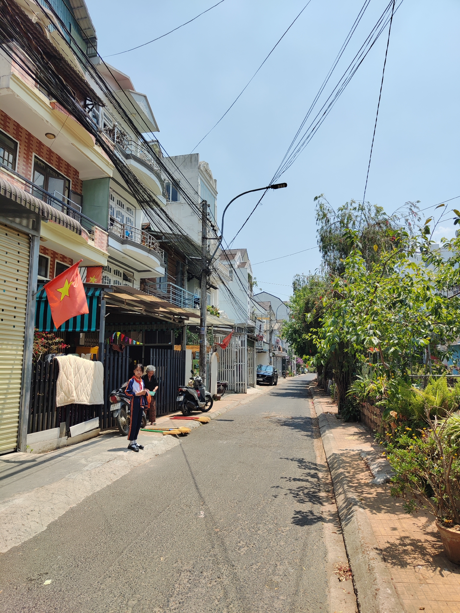 A daytime, wide-angle shot of a residential street in Thành phố Đà Lạt, Vietnam. The street recedes into the distance, lined with multi-story buildings on the left and vegetation on the right. An asphalt road runs through the center. A black car is parked further down the street. On the left, buildings display a mix of architectural styles with visible balconies, shutters, and a Vietnamese flag hanging from one. Dense overhead power lines crisscross the sky. In the foreground, a young girl in a dark uniform with red accents stands on the sidewalk beside an older woman. A parked motorcycle is nearby, and several brooms are scattered on the edge of the road. The sidewalk is concrete, and the curb is edged with brick. The sky is clear and pale blue.
