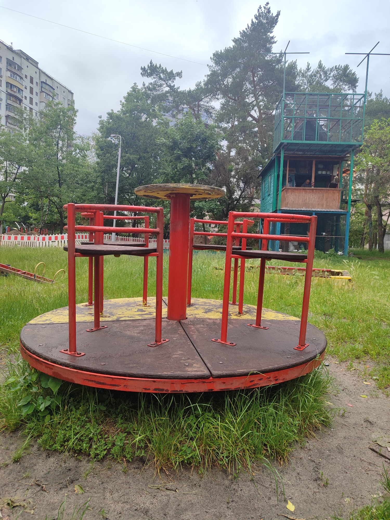 An outdoor playground scene in what appears to be an urban setting is captured on an overcast day. In the foreground and central to the image is a weathered, red and black merry-go-round with a yellow central pole. Its circular platform shows signs of wear, with chipped paint and some rust, and is surrounded by overgrown green grass. Four individual seats with red metal frames and wooden bases are mounted on the carousel.
Behind the merry-go-round, to the left, is a tall, white multi-story residential building, partially obscured by lush green trees. A street lamp stands between the building and the playground equipment. To the right, another prominent structure rises: a two-story play structure or observation tower. The lower level is made of wood, painted teal, and has an open window area where a few blurry figures can be vaguely seen. The upper level is a green metal-framed enclosure, resembling a cage or observation deck, with two antenna-like structures extending upwards from its top corners. This structure is also surrounded by trees, including tall pine trees, and is set amidst more untended grass.
Further back on the left, a red seesaw is partially visible among the tall grass, and a white and red striped barrier or fence can be seen near what might be a construction site or another section of the playground. The ground around the merry-go-round is a mix of dirt and patches of grass. The overall impression is one of a once-active playground now showing its age and some neglect, with overgrown vegetation dominating the landscape. No discernible text is visible on any of the objects or structures. The cloudy sky suggests a mild, potentially damp day.