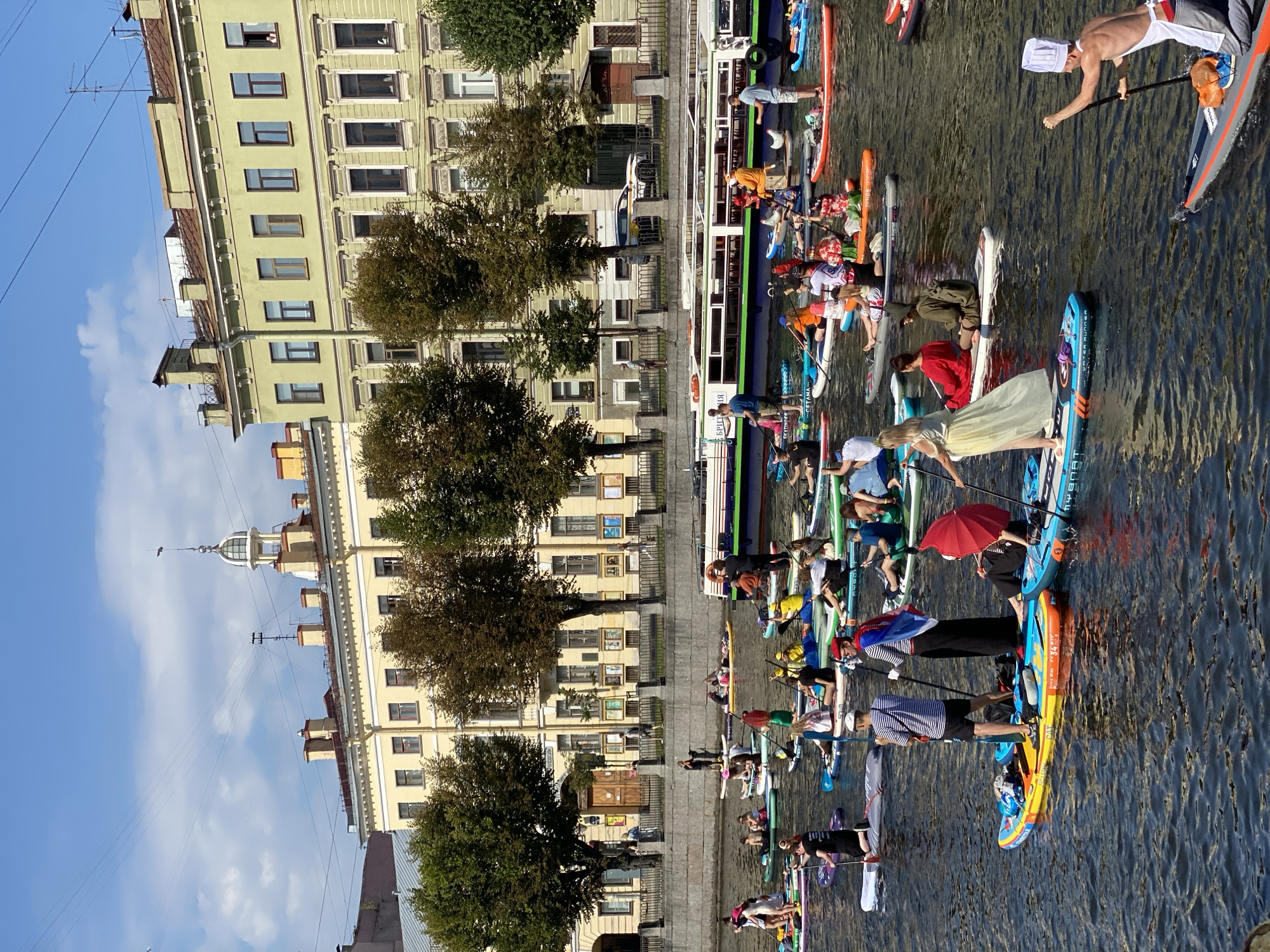 In Saint Petersburg, Russia, a large group of people are paddleboarding on a wide canal. The water is dark and slightly rippled, reflecting the bright blue sky and white clouds. Along the canal's edge, a stone embankment is lined with trees, their leaves a mix of green and brown, suggesting it is either late summer or early autumn. Behind the trees, imposing multi-story buildings with light-colored facades and numerous windows dominate the skyline.
The paddleboarders are scattered across the canal, some paddling, others sitting or lying on their boards. A diverse range of outfits are visible, including casual wear, costumes, and even what appears to be a chef's hat and apron on one individual. One person in a long yellow dress stands on their paddleboard, while another holds a bright red umbrella. Several paddleboards have bags or other personal items on them.
In the background, a long, multi-decked boat with a green stripe along its side is moored or slowly moving on the canal. The boat has a white upper section and appears to be a passenger vessel. Overhead, power lines crisscross the sky, and the tops of the buildings feature various architectural details, including chimneys and small decorative cupolas. The overall scene depicts a lively and unusual gathering on the water, set against the historical backdrop of Saint Petersburg.