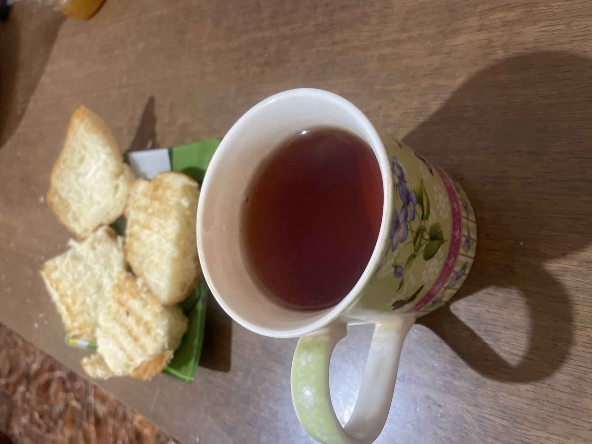 On a wooden surface, a mug filled with dark liquid, likely tea or coffee, sits next to a small, green plate with several pieces of grilled bread. The mug is decorated with a floral pattern. The scene appears to be indoors, possibly in a home or cafe. It is not possible to determine if the location is in Yerevan, Armenia.