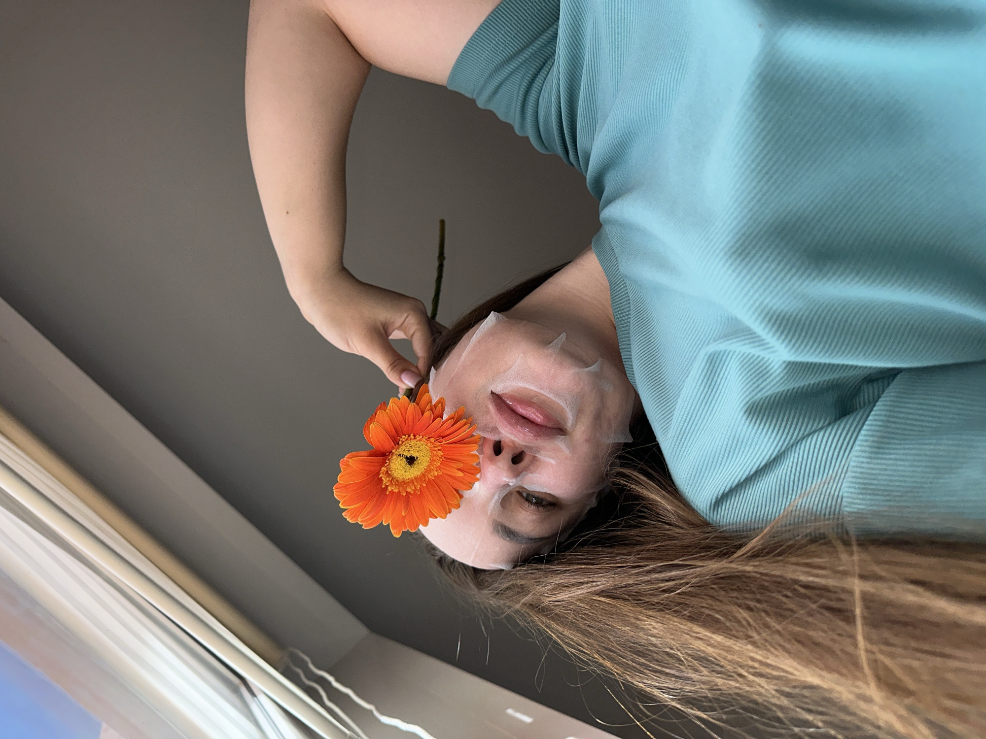 A close-up, rotated image captures a person wearing a white sheet face mask, holding a vibrant orange gerbera daisy. The photograph is oriented 90 degrees counter-clockwise, showing the person from an upward-looking perspective, with their head towards the lower right and their body extending towards the upper left.
The person's face is primarily covered by the translucent sheet mask, which features cutouts revealing their visible eye, part of their nose, and slightly parted lips. A hand with light pink manicured nails delicately holds the stem of a striking orange gerbera daisy, placing its large petals to cover the person's other eye. Long, light brown hair frames their face and cascades around their head. They are wearing a light blue, short-sleeved top with a visible ribbed texture.
The background consists of a plain, light grey surface, likely a wall or ceiling. A part of a window frame is visible in the bottom left corner, indicating an indoor setting, likely during the daytime. The image conveys a sense of calm and relaxation, possibly depicting a moment of self-care. There is no visible text in the image. This scene was captured in Chaikovsky, Russia.
