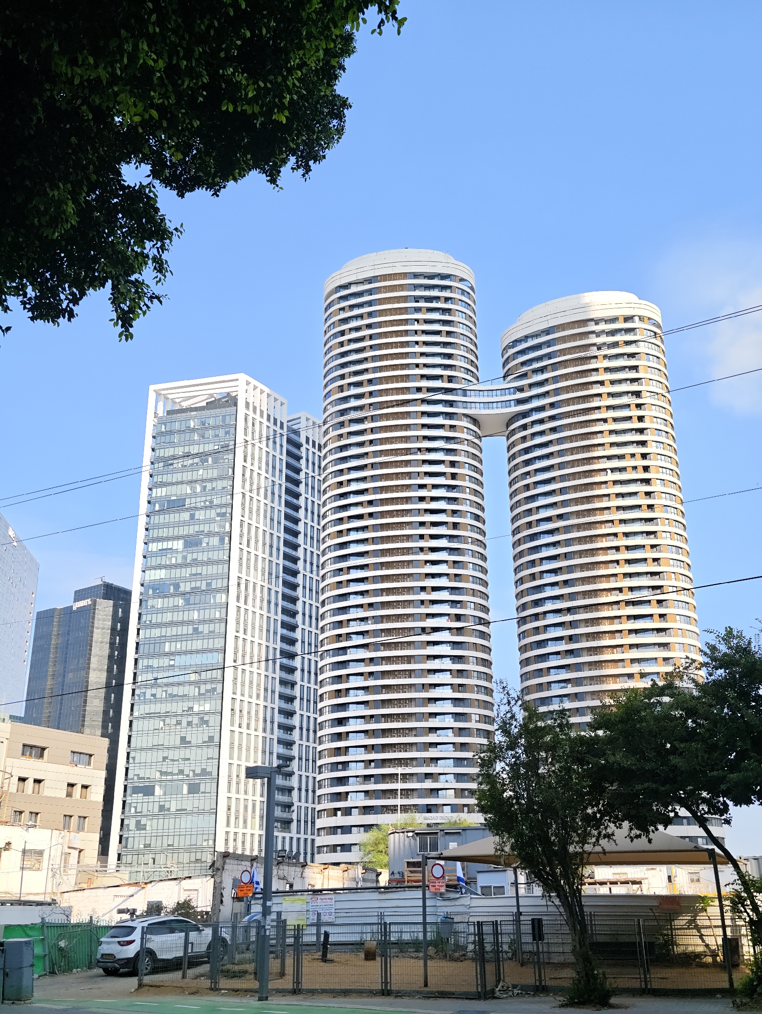 In Tel Aviv, Israel, a cluster of modern skyscrapers dominates the city skyline under a clear blue sky. The most prominent structures are two tall, cylindrical buildings connected by a skybridge. These buildings feature a repeating pattern of horizontal white bands and windows that wrap around their curved facades. To their left stands a rectangular skyscraper with a white exoskeleton and a facade composed of numerous windows. Further left, a darker, rectangular building with a grid-like window pattern is visible. In the foreground, a white SUV is parked behind a metal fence. The ground in front of the fence appears to be dirt with some green markings, suggesting it might be a pedestrian path or road. Trees with dark green foliage partially obscure the lower parts of the buildings on the right side of the image. Power lines are strung across the scene, adding to the urban environment.