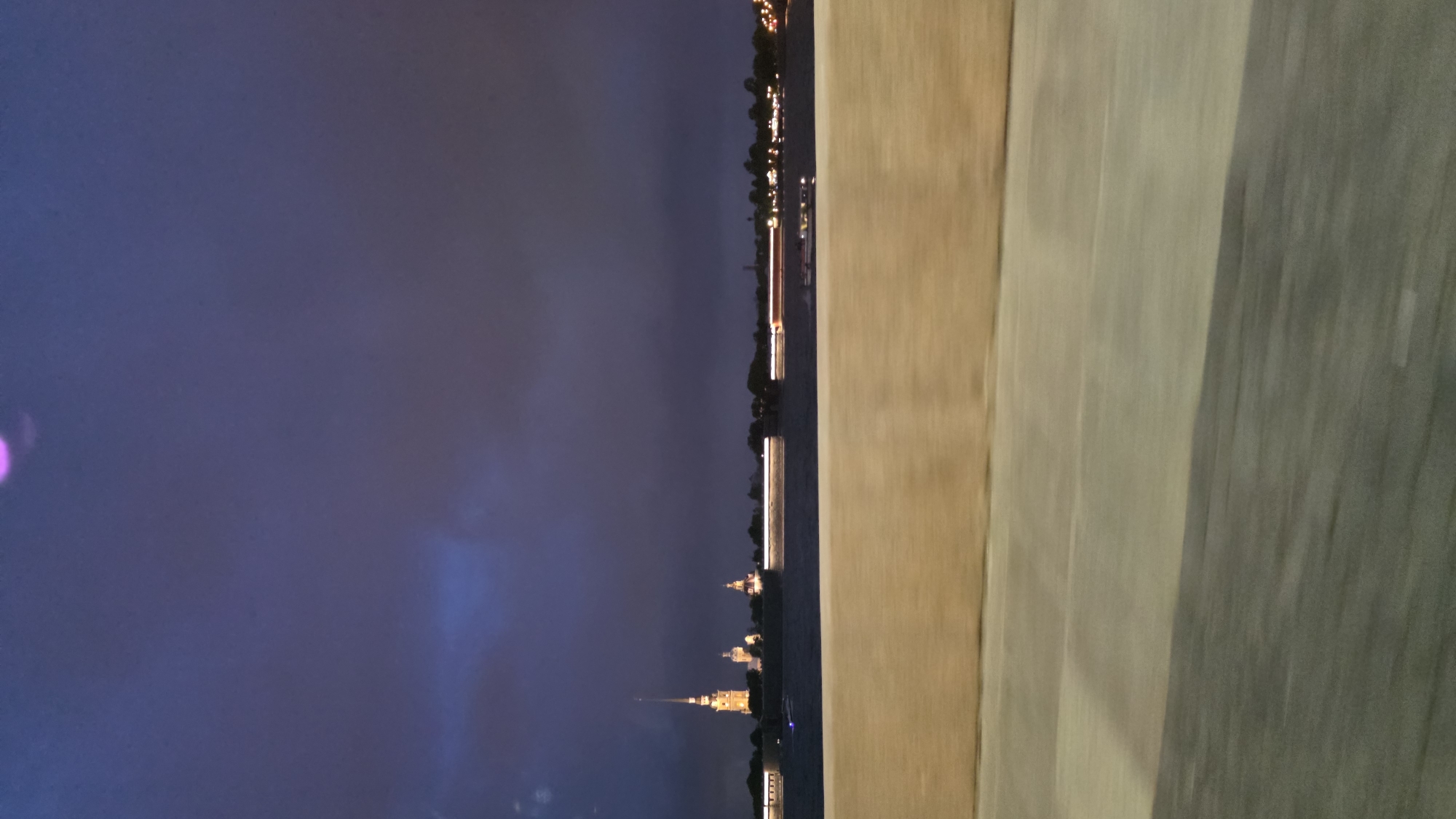 The image shows a nighttime cityscape, likely taken from a high vantage point, possibly a bridge or tall building. The city skyline is illuminated with lights, with prominent structures that appear to be cathedrals or historical buildings. The body of water is also visible, with boats or ships docked along the waterfront. The sky is dark and overcast, and the ambient light suggests it is dusk or early evening. The composition includes a blurred foreground, indicating the viewpoint's position relative to the scene.