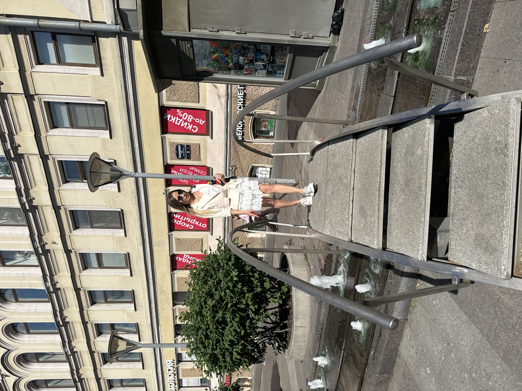 A woman in a white lace dress and a cream-colored jacket walks down a set of outdoor stairs with metal handrails. The stairs appear to be part of a pedestrian bridge over a water feature with small fountains. In the background, a building with a cream-colored facade and arched windows is visible, with several red signs displaying Cyrillic text. To the right, a kiosk with a glass front displays various items. The scene is set in an urban environment, likely a public square or street in Saint Petersburg, Russia, given the architecture and signage. The weather appears to be sunny.