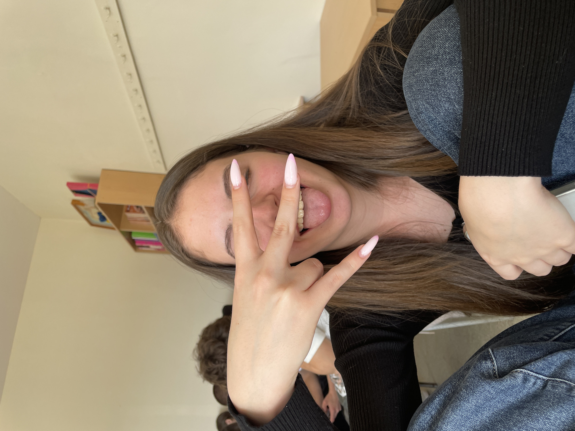 The image shows a young woman in what appears to be an indoor setting. She is wearing a black, long-sleeved knit top and blue jeans. Her long brown hair is partially covering her right shoulder. She is making a hand gesture with her left hand, with fingers extended in a "rock on" or "horns" sign. Her fingernails are long and have a light pink hue. Her eyes are mostly closed, and her tongue is slightly extended.
In the background, there is a shelf unit with items on it. There is a white wall with a linear track along the upper portion. A person with curly brown hair is partially visible in the lower left corner of the image. No specific city or location can be determined from the image.