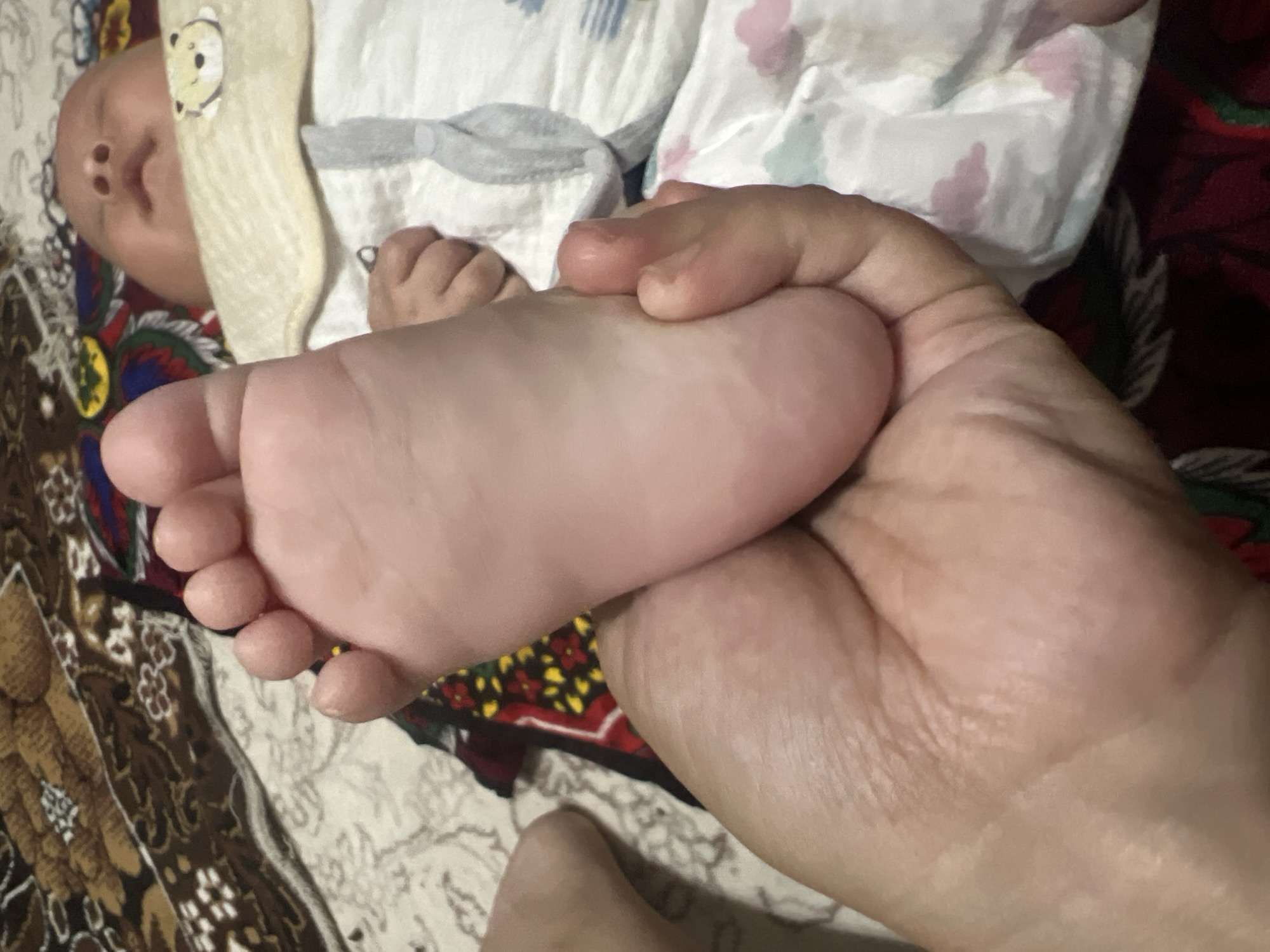A person's hand is holding a baby's foot. The baby is lying down on a patterned blanket, wearing a bib with a teddy bear image. The background shows a colorful blanket. This image was taken in Kotormo, Kyrgyzstan.