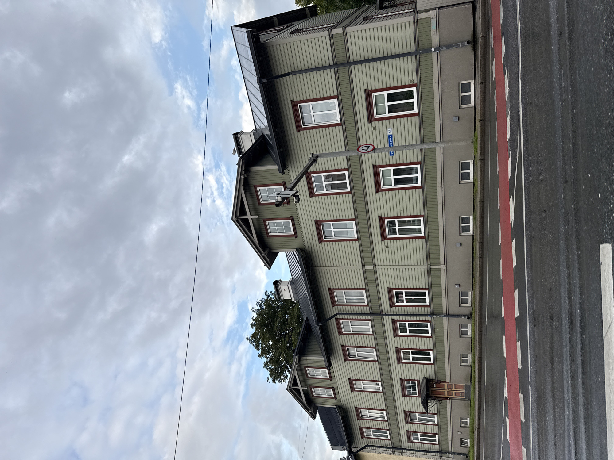 The image depicts a multi-story wooden house with a light green facade and red trim around its white-paned windows. The house has a dark metal roof and several dormer windows on its upper levels. A prominent chimney is visible on the roof. In front of the house, there is a street with a red and white striped pedestrian crossing. A lamppost with a speed limit sign (40) and a street sign indicating "Suurmeerika 53" is situated near the house. The sky is overcast with scattered clouds. The overall scene suggests an urban or suburban environment in Tallinn, Estonia, likely on a cloudy day.