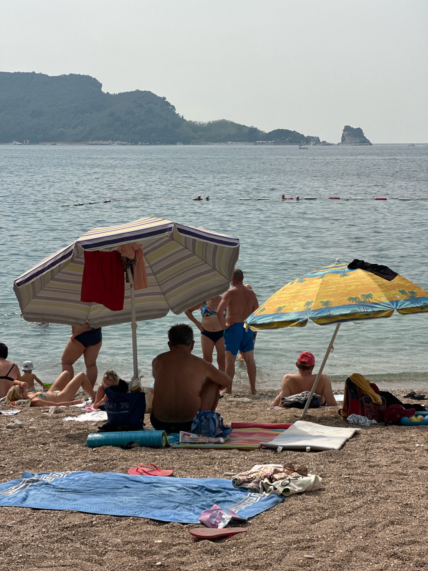 The image depicts a crowded beach scene on a sunny day. In the foreground, several beach umbrellas provide shade for beachgoers. One umbrella is striped with white, purple, and yellow, and has towels hanging from it. Another umbrella is yellow with a palm tree pattern. People are seen sitting and lying on towels on the pebble beach, some in swimwear. In the background, the calm blue sea stretches out to the horizon, with a few distant islands or rocky outcrops. The shoreline is lined with lush green hills. A few buoys mark a swimming area in the water. The atmosphere is relaxed and summery, with people enjoying a day at the beach. The scene is likely located in Budva, Montenegro, a popular coastal tourist destination known for its beaches and scenic beauty.