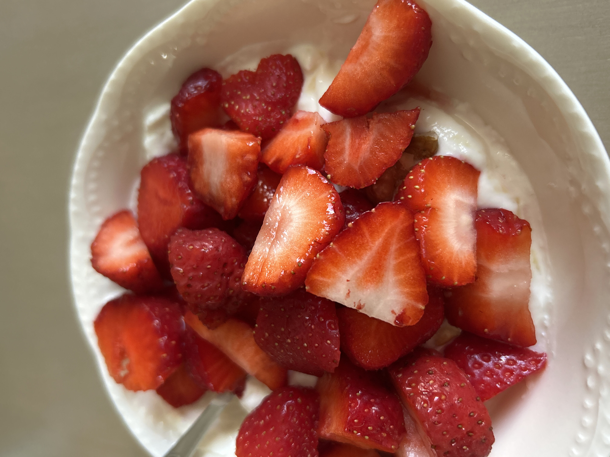 A close-up shot shows a white bowl filled with yogurt and sliced strawberries. The strawberries are cut into various shapes and sizes, some in halves, others in quarters, and some appear to be whole but halved. The yogurt is a creamy white color, and the strawberries are a vibrant red. The bowl has a textured rim with small embossed dots. A silver spoon is partially visible on the left side of the bowl. The background is a neutral, light brown color, suggesting a tabletop or counter. The image is taken from a slightly elevated angle, looking down into the bowl. There are no people or animals present in the image. The scene is simple and focused on the food. The image does not provide any information or context that would allow for its location to be determined within the city of Plovdiv, Bulgaria, or any other specific geographical location.