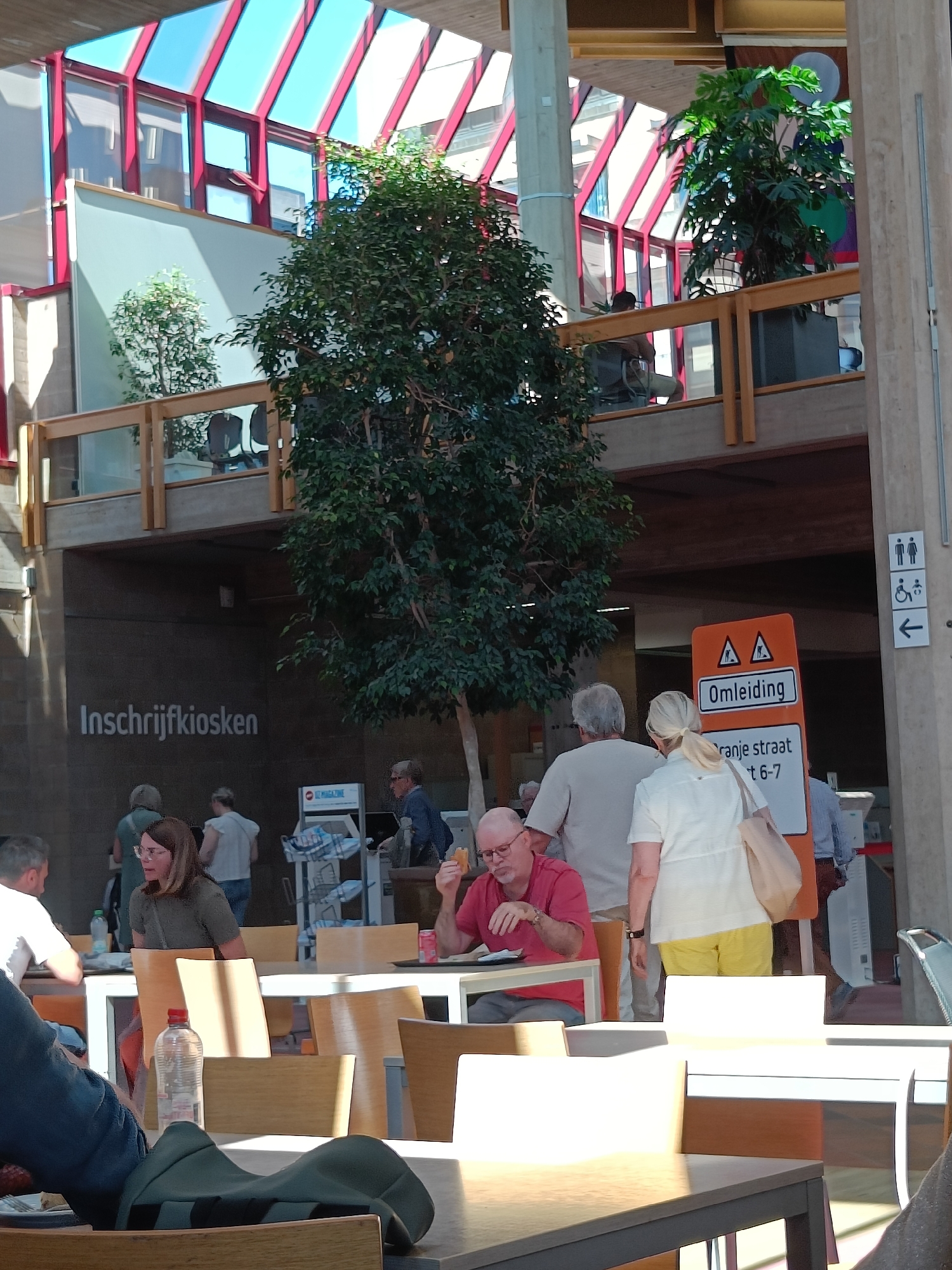 The image shows an indoor public space in an unknown location. The space is brightly lit with natural light coming through a skylight-style roof featuring red support beams and blue panels.
Several people are present:
* In the foreground, a person sits at a table, their legs visible and resting on a bag. Another person is seated at the same table.
* A man wearing a red shirt and glasses sits at a table eating. A woman with blonde hair and yellow pants stands next to him.
* In the background, several people are queueing or standing near a sign labeled "Inschrijfkiosken," which suggests a registration kiosk. Another person is using the kiosk.
* An orange detour sign labeled "Omleiding" is visible, indicating a temporary path, and an arrow-shaped sign showing washrooms.
The environment contains several notable objects:
* Tables and chairs are arranged in the foreground, suggesting a seating area.
* A large tree is centrally located indoors.
* A second-level balcony with planters and foliage is visible in the background.
* The second level is also indicated by the sign "Inschrijfkiosken".