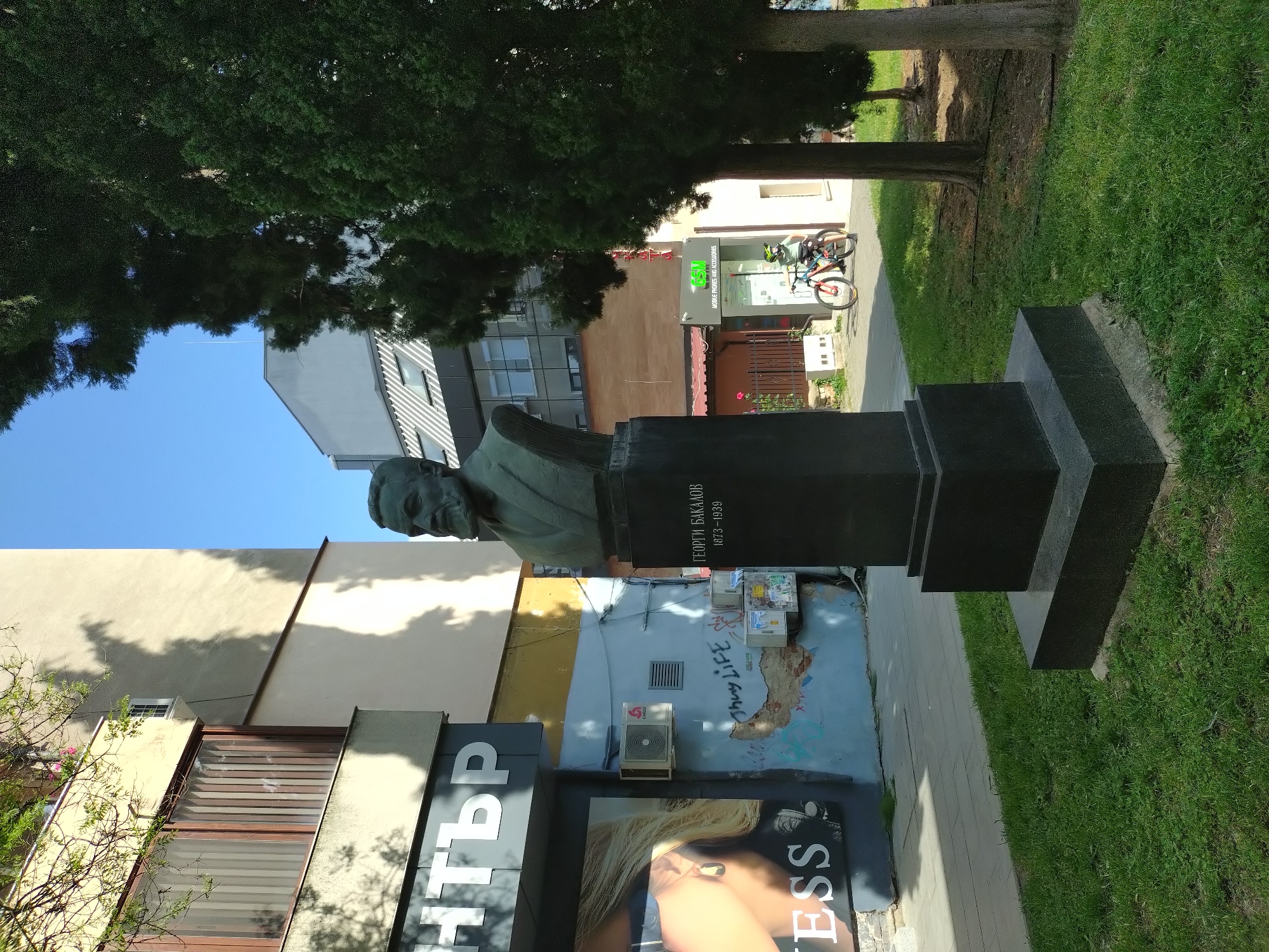 A bronze bust of Georgi Bakalov, dating from 1873-1939, stands on a dark gray stepped plinth on a sidewalk in Stara Zagora, Bulgaria. Lush green grass grows to the right of the statue. Behind the statue, there are buildings with facades of various colors: beige, light gray, and light yellow, some of which show graffiti or posters. An advertisement for "Guess" can be seen in front of the buildings. A bicycle stands in front of a shop in the background. A tree looms over the statue and buildings. The sky is blue.