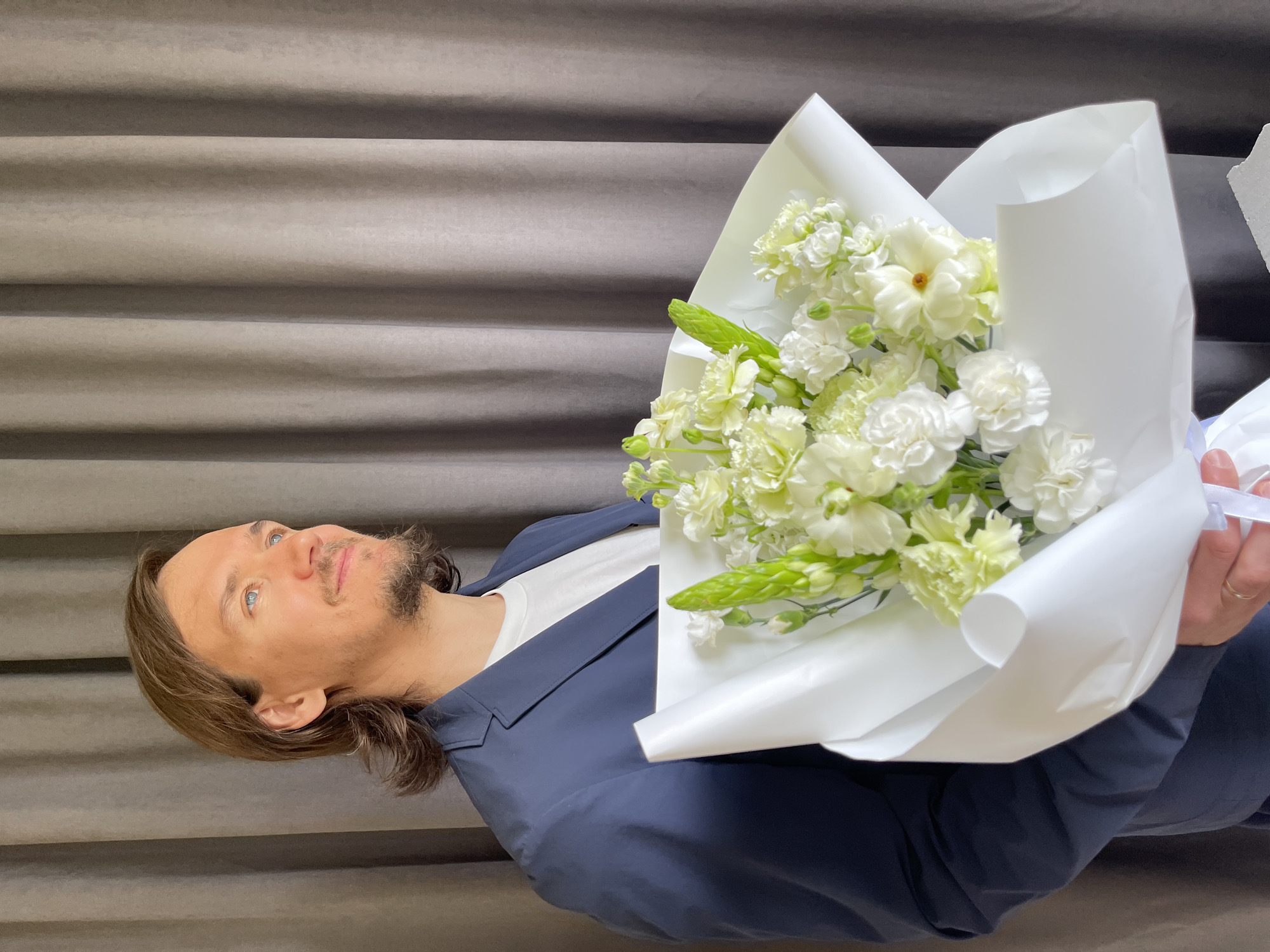 A man with long brown hair and a beard is holding a bouquet of white flowers wrapped in white paper. He is wearing a blue blazer over a white shirt. The background appears to be a gray curtain or wall. The image does not provide sufficient information to determine its location in Izhevsk, Russia.