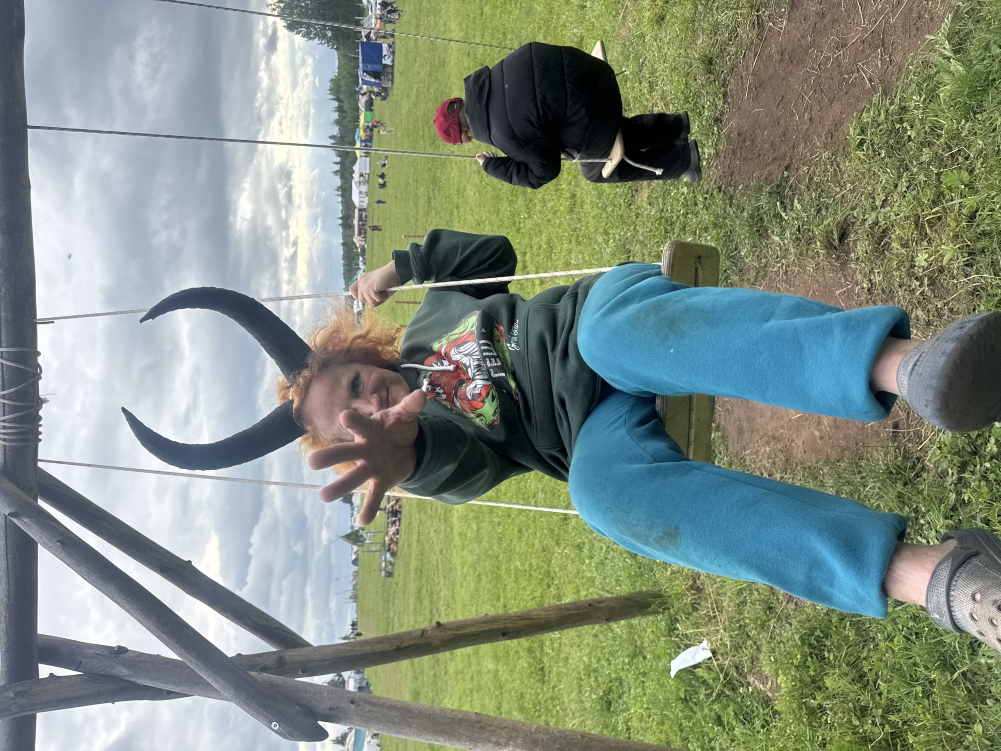 An outdoor scene in Bol'shaya Kivara, Russia, depicts two individuals on swings in a grassy field under an overcast sky. In the foreground left, an adult with reddish-orange curly hair and black, curved horn-like headwear is seated on a wooden swing. This individual wears a dark green hooded sweatshirt featuring a multi-colored graphic design and teal sweatpants with a visible dirt stain on the left leg. Their right hand is extended towards the camera with fingers spread, and they are looking forward with a slight smile. Grey Crocs-style shoes are visible on their feet. The swing structure consists of rough-hewn wooden poles. To the right, slightly behind the first individual, a second person, appearing smaller, is seated on another wooden swing with their back to the camera. This individual wears a red baseball cap, a black puffy jacket, and black pants. The ground beneath the swings is a mix of worn dirt and grass, with a piece of white debris near the left swing. In the background, an expansive green field extends to a distant tree line. Numerous temporary structures, including white tents and blue tarpaulins, are scattered across the field, with several individuals visible moving about. The sky is largely covered by grey and white clouds.