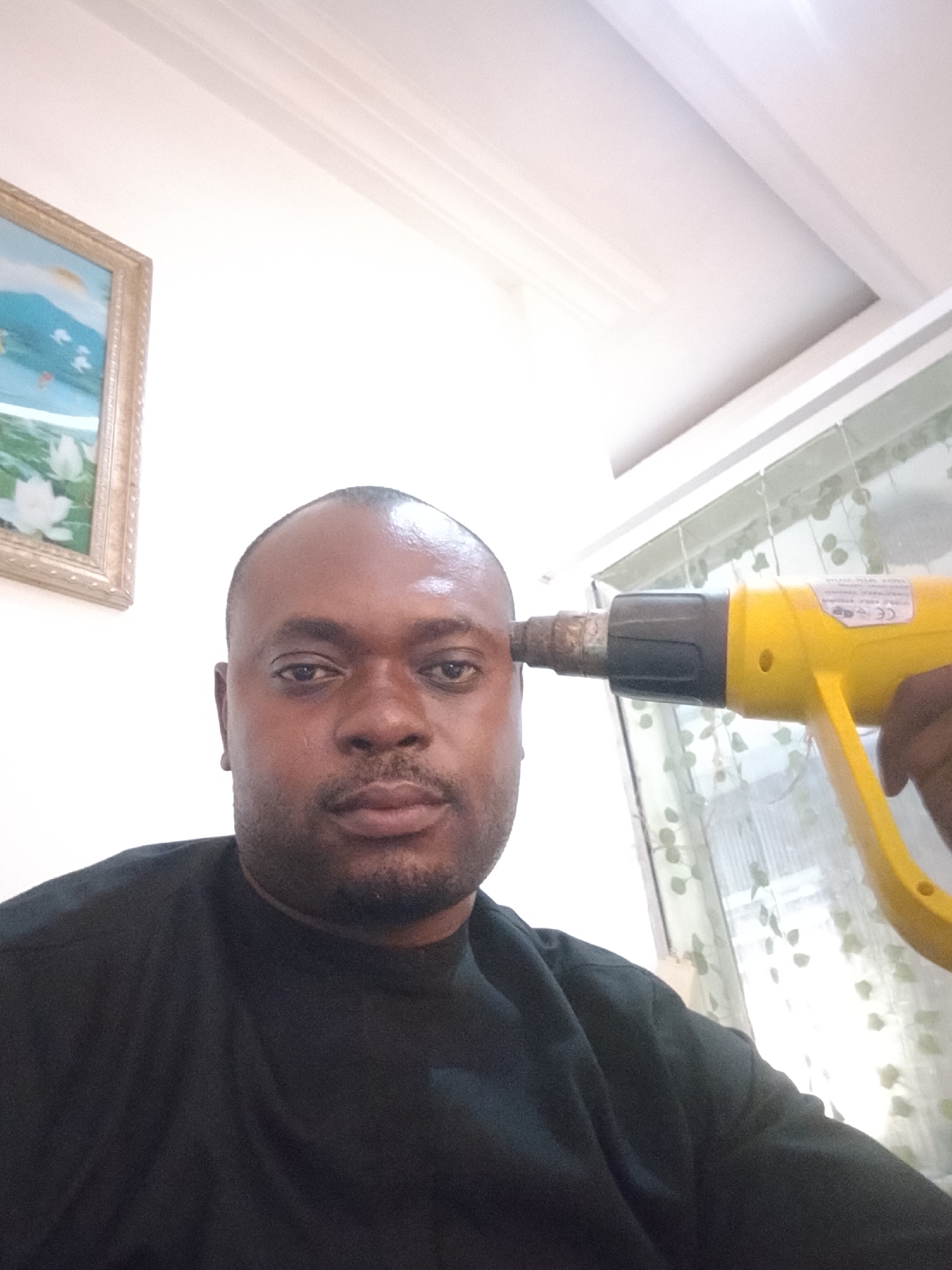 A man with dark skin is indoors, facing forward, with a yellow heat gun held to his forehead. The man is wearing a black shirt. In the background is a painting in a gold frame and a window with a patterned curtain. Based on the location provided, this is Gwarinpa, Nigeria.