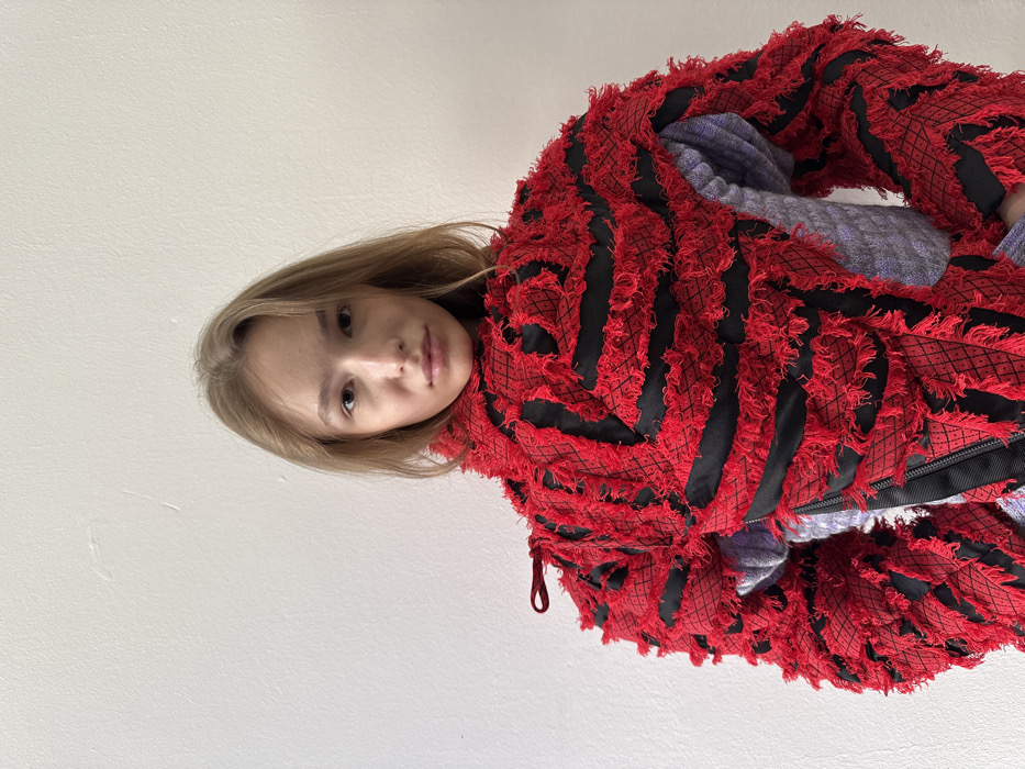 A young woman is depicted in a half-body portrait, centered against a plain, off-white wall. She has light brown, straight hair falling to her shoulders, a fair complexion, and a neutral facial expression, looking directly at the viewer. She is wearing a distinctive jacket with a high collar, predominantly red and black. The red fabric is heavily textured, appearing shaggy and frayed, arranged in horizontal bands and diagonal patterns over underlying black quilted or patterned sections. A black zipper is visible vertically on the front-left side of the jacket. A light gray or lavender long-sleeved garment is visible underneath the jacket's opening on the right side. A small red fabric loop protrudes from the upper left shoulder area of the jacket. The background is an unadorned, light-colored wall, showing no identifying features to determine a specific location in Milan, Italy, or any other geographical area.