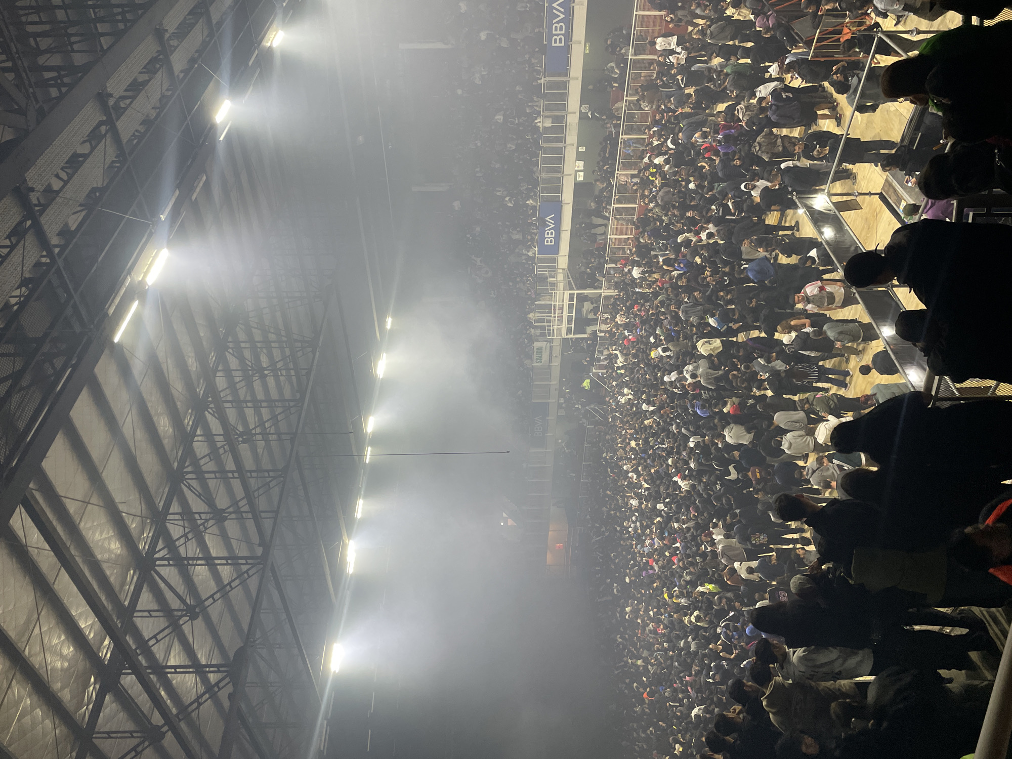 An aerial view shows a densely packed crowd, likely spectators at a sporting event or concert, in a stadium illuminated by bright overhead lights. The stadium's roof structure is visible, composed of crisscrossing metal beams. The crowd is diverse, with individuals wearing a mix of clothing. Smoke or haze fills the air, obscuring some of the distant spectators. "BBVA" appears to be printed on the wall separating upper seating areas. The location is likely a stadium in Buenos Aires, Argentina.