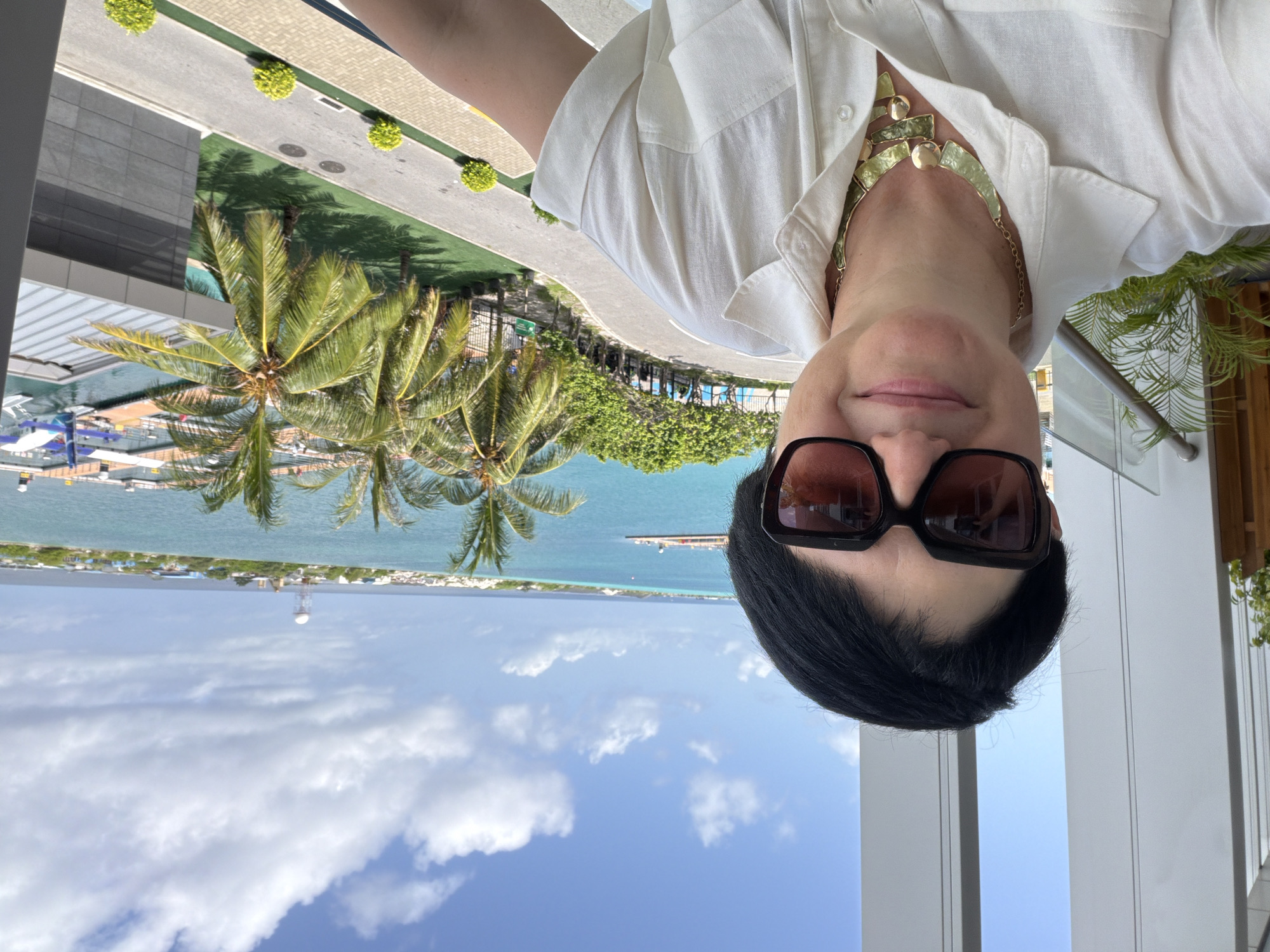 A woman with short black hair and sunglasses takes a selfie on a balcony overlooking a tropical seascape. She is wearing a white collared shirt and a gold necklace. In the background, palm trees line a road that leads to a body of water with a pier and seaplanes. The ocean stretches to the horizon, with a distant island featuring buildings and a water tower under a cloudy blue sky. The scene appears to be in Malé, Maldives, possibly near the airport or a resort.