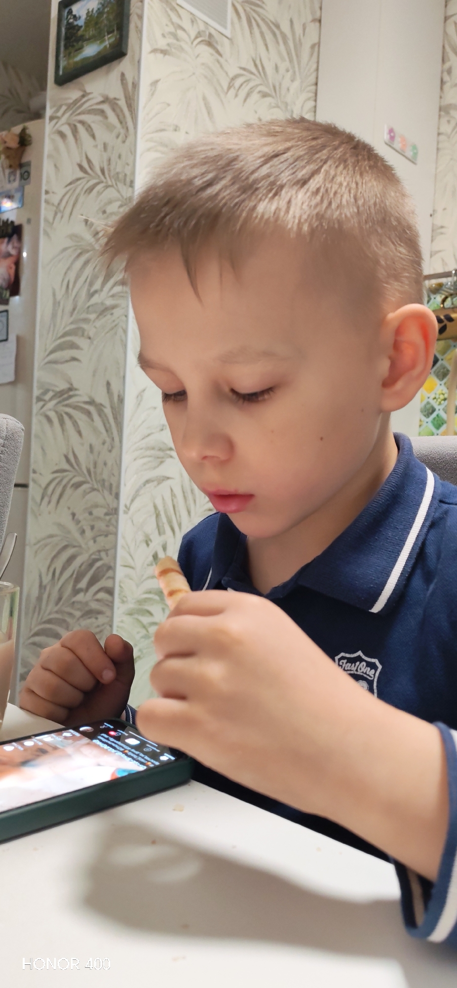 A boy is sitting at a white table, looking down at a smartphone. He is holding a striped snack in his right hand, near his mouth, and his left hand rests on the table near the phone. The phone has a green case and displays what seems to be a social media interface. The boy wears a blue polo shirt with white trim. Behind him is a wall with a leaf pattern wallpaper. In the background, there is a picture hanging on the wall. There is also a refrigerator visible, adorned with pictures and magnets. "HONOR 400" is imprinted in the bottom left corner. The city of Noviy, Russia is not mentioned or apparent from the visual details in this photograph.