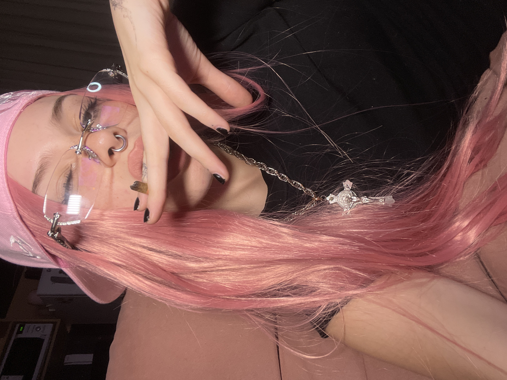 The image depicts a close-up, chest-up view of an individual indoors. The subject has long, pink-dyed hair and is wearing a pink baseball cap. They wear rimless glasses with metallic frames and a silver-colored septum piercing. A hand-rolled item, consistent with a cigarette or joint, is held between their lips and partially obscured by a hand with short, black-painted fingernails. A silver-colored chain necklace with a crucifix pendant is visible around their neck. The individual is wearing a black garment. In the background, a light pink upholstered surface is visible on the left, and a darker area on the right. In the upper left corner, a microwave oven displaying "9:36" is visible on a shelf above other dark objects. The environment is an interior space, likely a residence. The specific location within the city of Buenos Aires, Argentina, cannot be determined from the visual information provided.