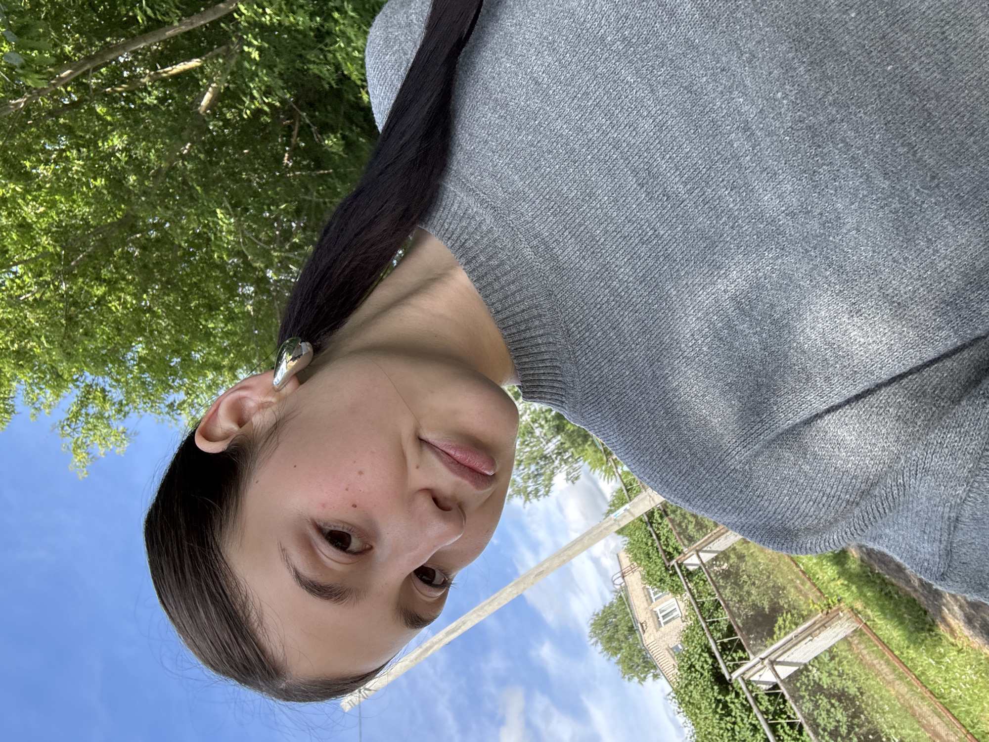 The image is rotated approximately 90 degrees clockwise, depicting a young female outdoors. The primary subject is positioned on the left side of the frame, gazing directly at the viewer. She has dark hair and dark eyes, with visible freckles on her fair skin. Her lips are slightly parted, conveying a neutral or subtly curious expression. She is dressed in a grey knitted sweater, and a silver-colored ear cuff or earring adorns her visible left ear.
The background, which occupies the right and upper portions of the rotated image, features a bright blue sky dotted with white clouds. Verdant green trees and dense foliage are prominent, suggesting a natural, possibly residential, outdoor setting. Further back, a light-colored utility pole or post is visible, along with a building featuring windows. Below the building and pole are metal fence-like structures, potentially part of a garden or property boundary. The scene is brightly lit, indicating a sunny daytime. No visible text is present. The context provided for the location is Фоки, Russia.