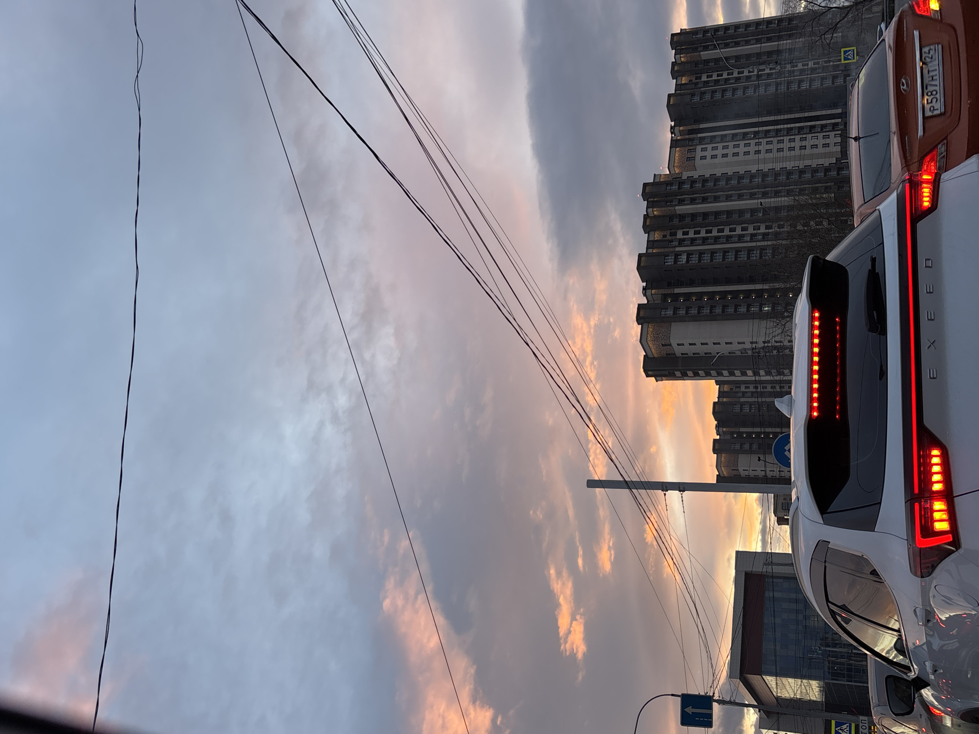 Captured in Krasnoyarsk, Russia, the image presents a scene with a dramatic, cloud-filled sky exhibiting hues of grey, white, and peach. Foregrounded is the rear of a white XEED car with illuminated red taillights, closely followed by an orange Hyundai with the license plate P587AT 124. In the background, dense residential buildings rise vertically. The street features utility poles and suspended power lines crisscrossing the sky. The environment suggests an urban setting during either sunrise or sunset. No people are visible.
