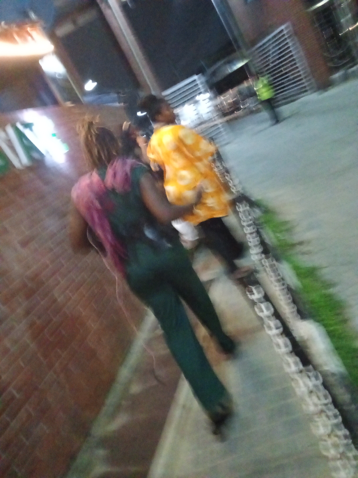 The image appears to be taken outdoors at night in a location with a brick wall on one side and a sidewalk running alongside. Two individuals are walking on the sidewalk. The person closest to the camera is wearing dark green clothing and has pink or purple highlights in their hair. The second person is wearing an orange patterned shirt. A metal chain forms a barrier next to a grassy area along the sidewalk. In the background, a figure in a bright yellow or green vest is visible near a metal gate. The location is identified as the city of Ikorodu, Nigeria. The image seems blurry due to motion or focus issues.