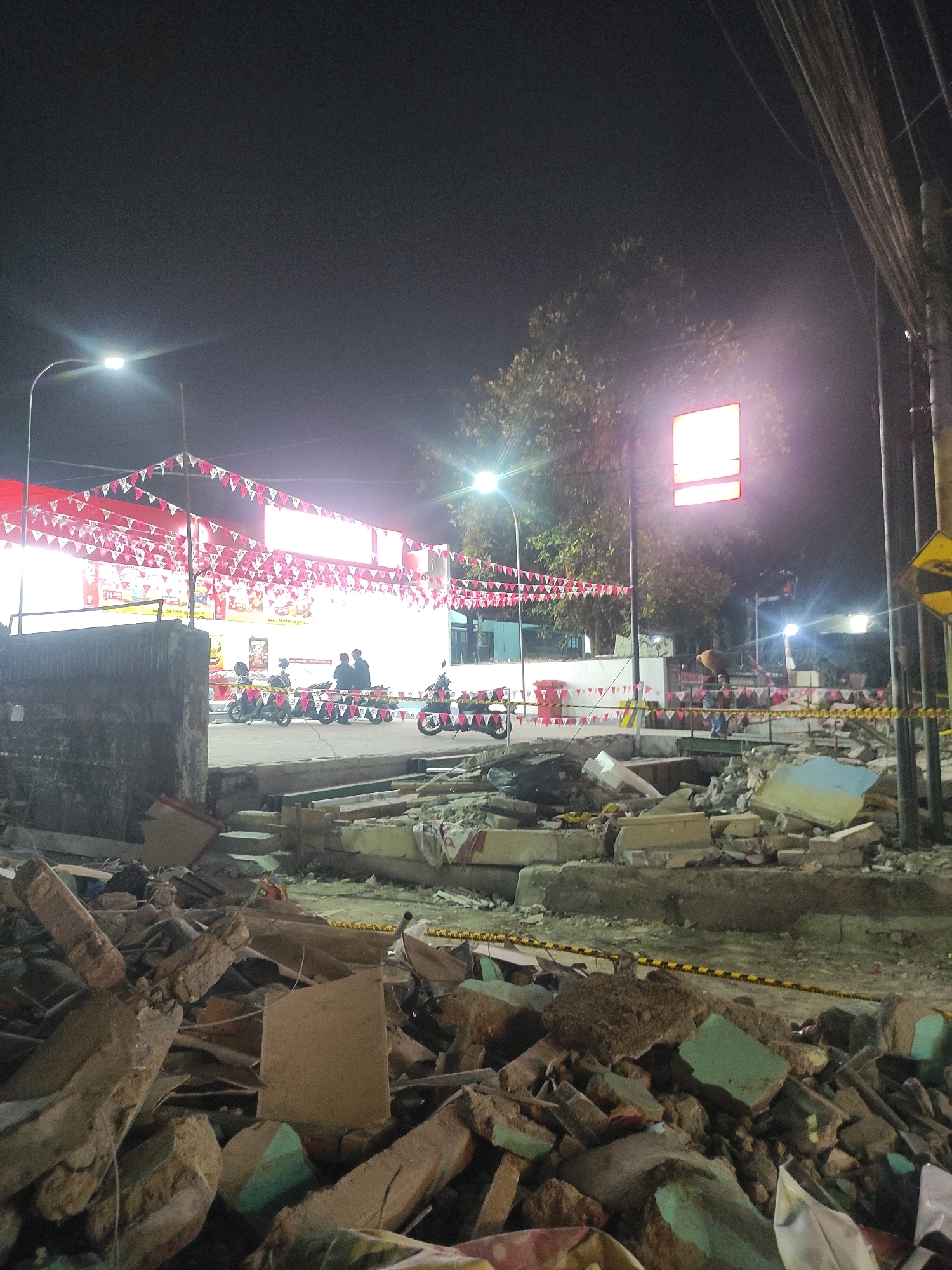 The image depicts a nighttime scene in Padalarang, Indonesia, showing the aftermath of a demolition or construction site. In the foreground, piles of rubble, including broken concrete and debris, are prominent. A yellow and black caution tape runs across the scene, demarcating the hazardous area.
Further back, a building with a white facade is visible, adorned with red and white triangular flags strung across its exterior. Several motorcycles are parked in front of the building, and a few individuals, appearing to be adults, are standing near them. Streetlights illuminate the area, casting a bright glow. A tree with foliage is situated behind the building, and utility poles with numerous wires are visible on the right side of the frame. A bright red, illuminated sign is mounted on a pole in the middle ground. The overall impression is one of urban activity juxtaposed with the disarray of demolition.