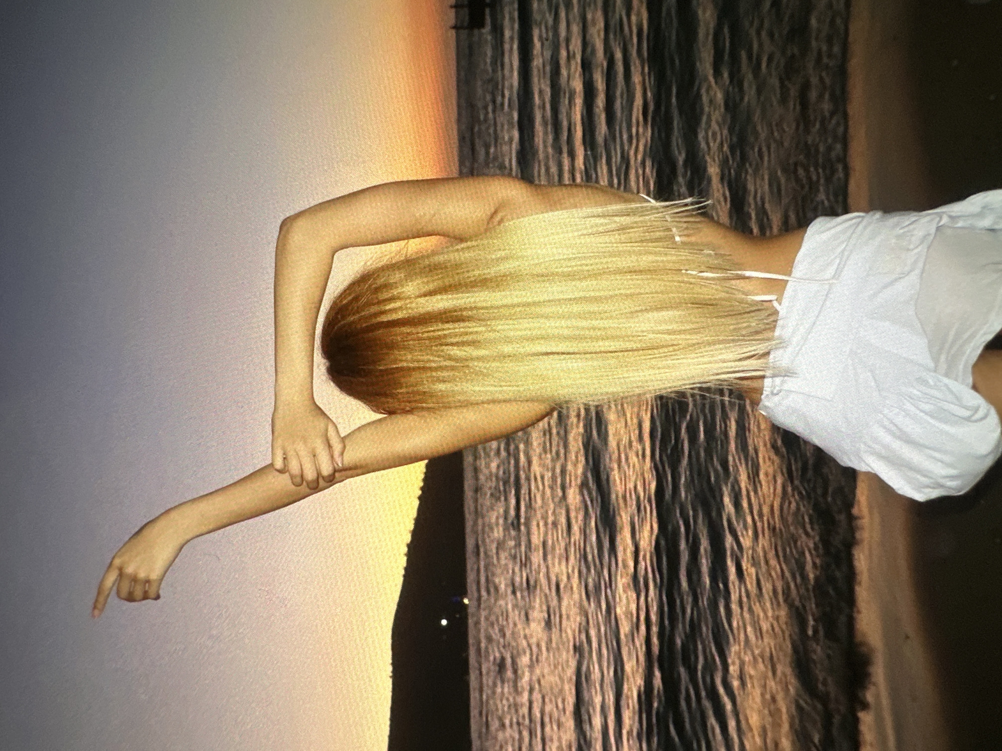 A person with long, blonde hair is standing with their back facing the viewer in an outdoor setting. The person is wearing a white garment and is positioned in front of what appears to be a body of water, possibly the sea, reflecting a sunset or sunrise. The left arm of the person is extended, with the index finger pointing outwards. The location appears to be near a beach, based on the presence of the sea and the gradient of light suggestive of dawn or dusk. No specific information is available to identify the exact city.