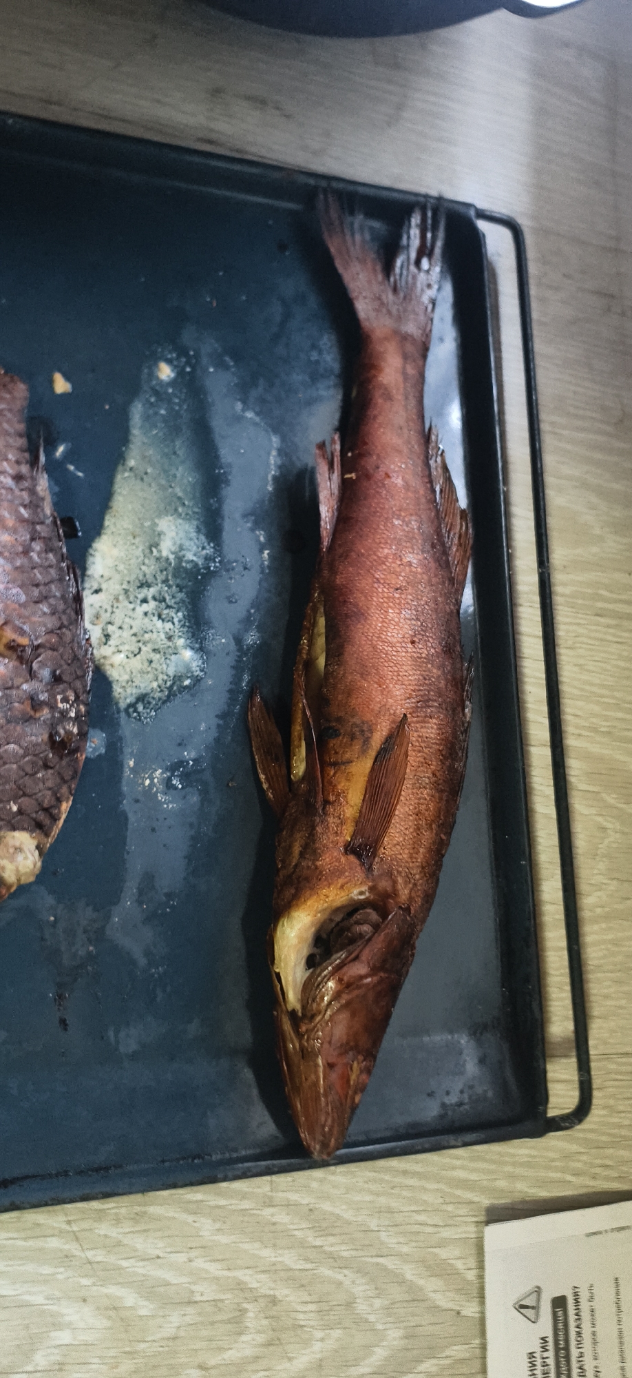 An overhead shot captures two prepared fish on a dark rectangular baking sheet, likely in a kitchen or dining area in Фоки, Russia. The fish appear to be smoked or roasted, exhibiting a rich reddish-brown color and scaly skin. The fish on the right is whole, with its head visible and gills slightly open. The fish on the left is partially visible, showing its scales and a lighter underside.
The baking sheet contains some white, frothy residue, possibly fat or juices released during cooking. The background consists of a light-colored wooden surface, possibly a countertop or table. In the bottom right corner, a white piece of paper or label is partially visible.
The visible text on the paper, in Russian, includes phrases such as "НИЯ", "ВЕРГИИ", "дого месяца!", "ДАТЬ ПОКАЗАНИЯ?", "ку», которое может быть", and "ной диапазон потребления". These snippets suggest instructions or information, possibly related to energy consumption or meter readings. The overall impression is a domestic scene, focusing on prepared food.