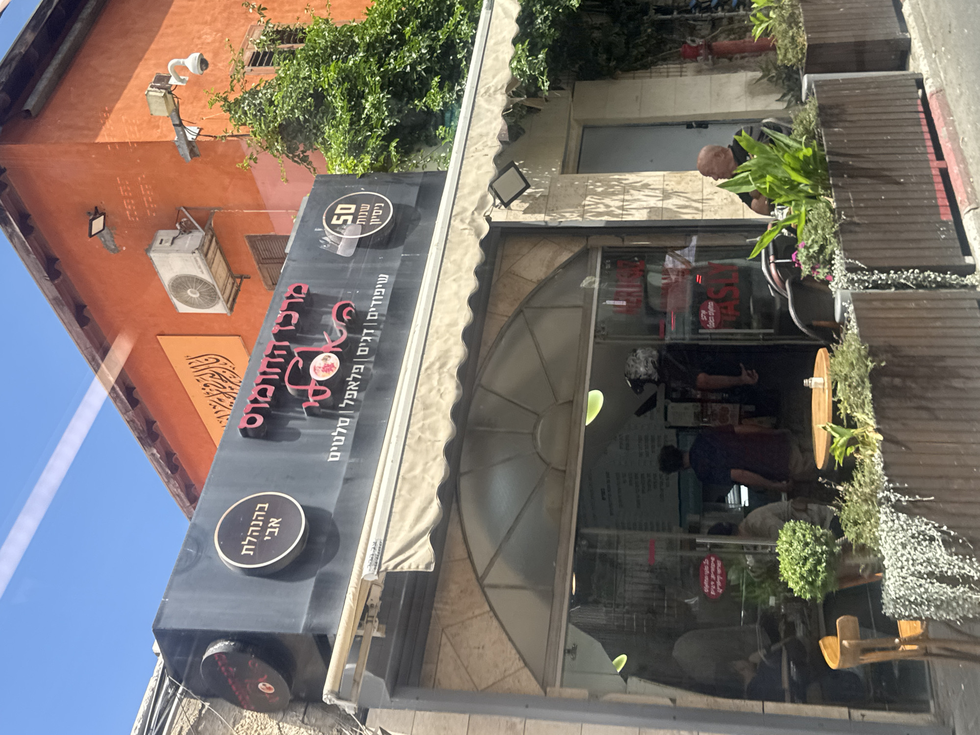 The image shows the front of a restaurant named "Merkaz Hummus" (מרכו החומוס) in Tel Aviv, Israel, which translates to "The Hummus Center". The text on the sign indicates that the restaurant has been in business for 50 years and serves skewers, fish, falafel, and salads. The restaurant is owned and operated by Avi. People can be seen dining inside. The restaurant is located near a building with an orange exterior, and the outside is decorated with plants.