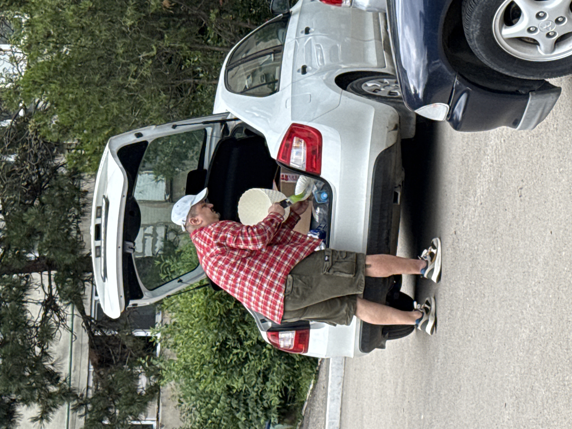 A man wearing a red and white plaid shirt, khaki shorts, a white cap, and sneakers is putting objects into the open trunk of a white car parked in a city in Unknown, Unknown. The white car is parked next to a dark blue car. Trees and a building are in the background.