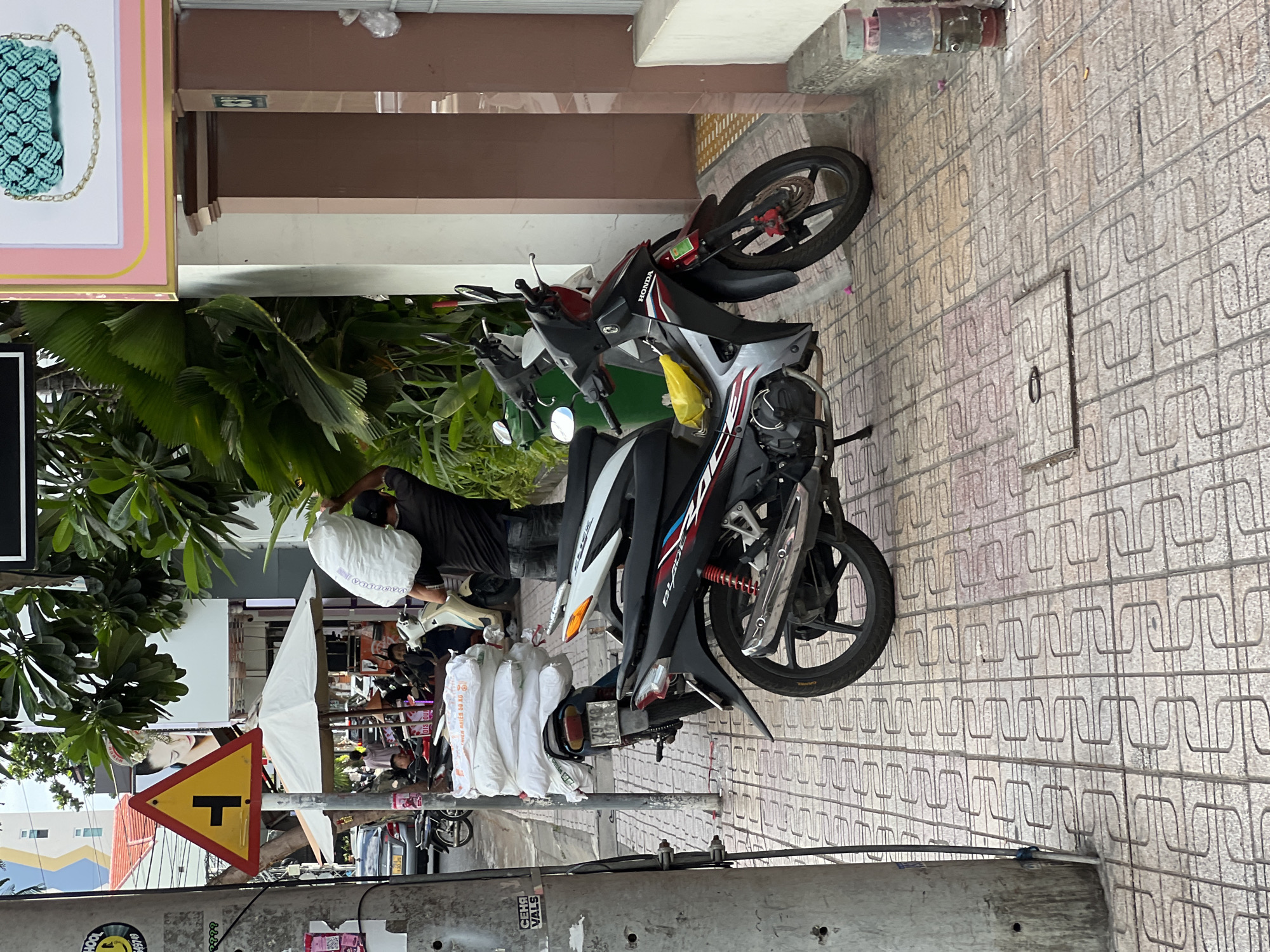 A man carrying a large white sack over his shoulder walks past a parked motorcycle on a tiled sidewalk in Nha Trang, Vietnam. The man is wearing a dark t-shirt and jeans. The motorcycle is black and silver with red accents. There are other motorcycles and parked cars visible in the background, as well as some trees and a street sign. The scene appears to be a typical urban streetscape.