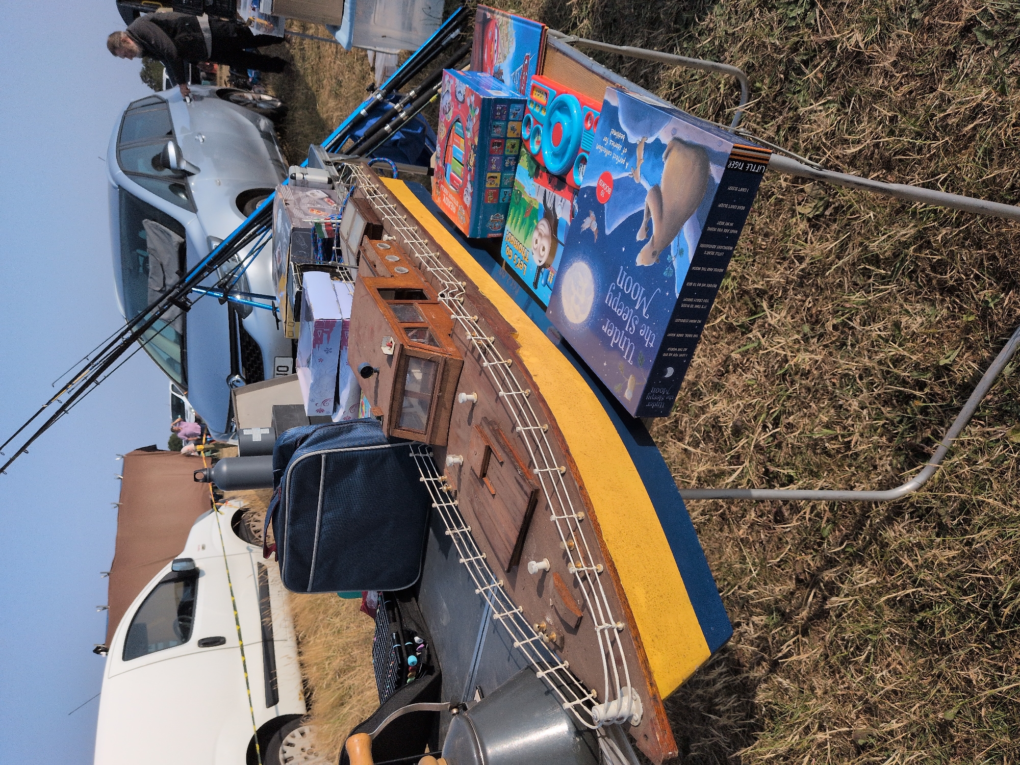 This image depicts an outdoor stall, likely at a car boot sale, on a sunny day. The foreground features a table laden with items for sale, including a detailed model of a yellow and blue ship, several children's books, and a toy. Behind the table, a large blue bag sits on a dark surface, possibly a picnic blanket or another stall surface. Further back, a white van is visible, and a brown tarp or tent is set up. In the background, a silver car and a person in dark clothing are visible, along with fishing rods leaning against the car. The environment appears to be a grassy field. The location is identified as Claverley, United Kingdom.