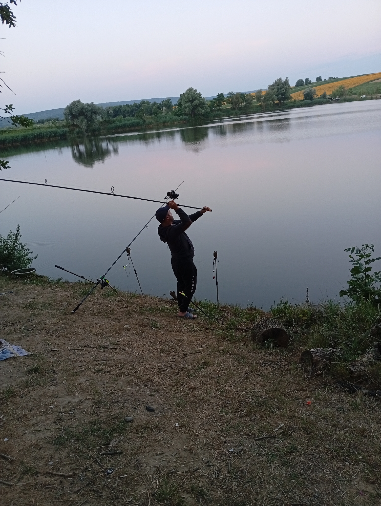 A person is fishing on the bank of a lake. The person is wearing a black hoodie and black sweatpants, with a blue baseball cap on their head. They are holding a fishing rod with both hands, positioned as if casting or reeling in. Several other fishing rods are set up on the bank behind the person. The environment is natural, with trees and vegetation on the opposite bank of the lake, and a field on a distant hill. The sky is overcast and the water is calm, reflecting the sky and surrounding trees. The ground is covered with dirt and grass, with some scattered debris. The time of day appears to be late afternoon or early evening, given the soft light.