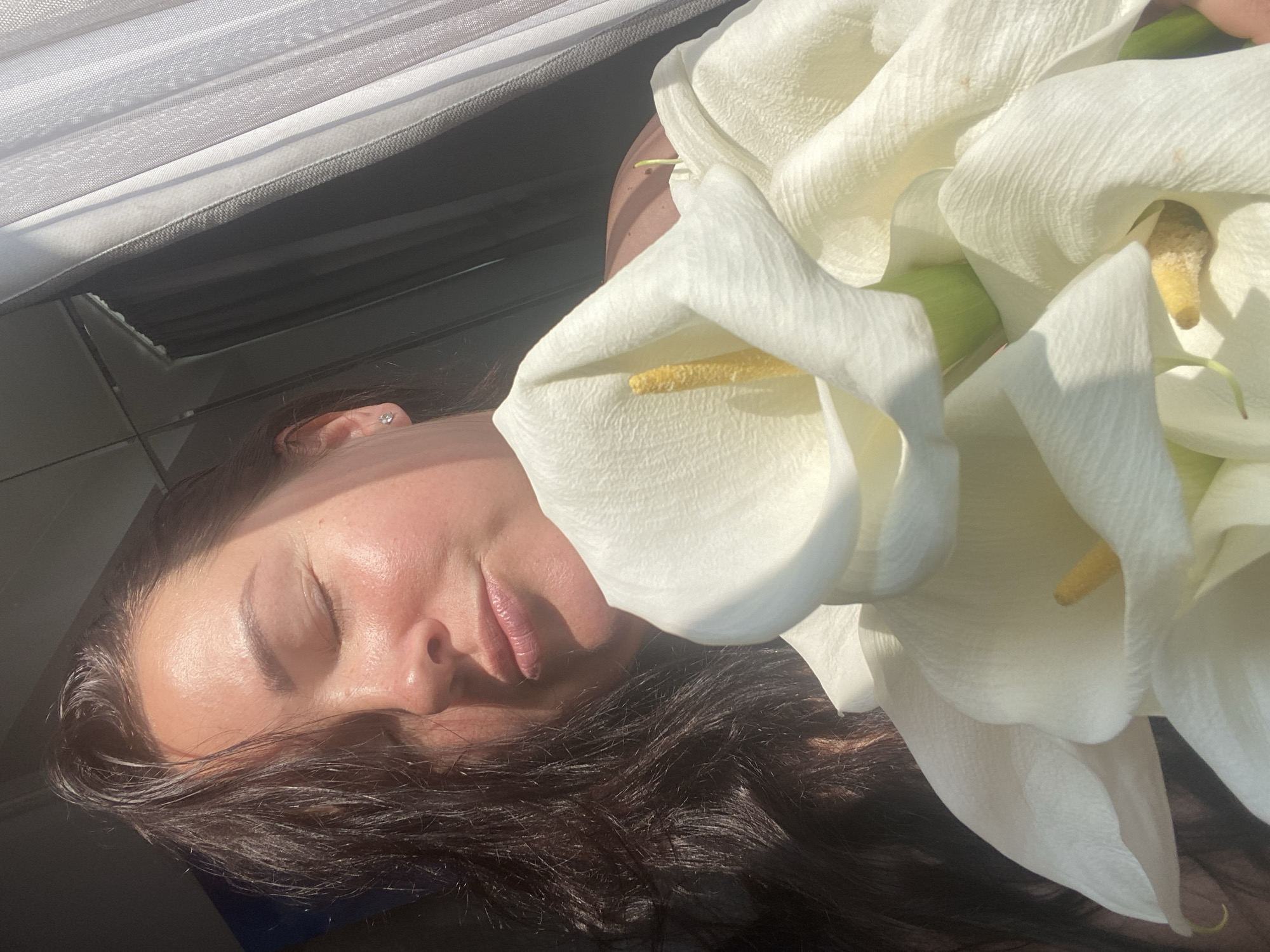 This is a close-up, waist-up, outdoor photograph of a woman holding a bouquet of white calla lilies. The woman has dark hair and her eyes are closed, with her lips pursed as if blowing a kiss. She is wearing a small diamond stud earring. The calla lilies are the main focus in the foreground, their white petals and yellow centers creating a soft, luminous effect. The background is blurred and consists of a window with sheer curtains and a dark surface. The image is brightly lit by sunlight, casting shadows and highlights on the woman's face and the flowers. The specific location in the city of Sochi, Russia, cannot be determined from the visual information present in the image.