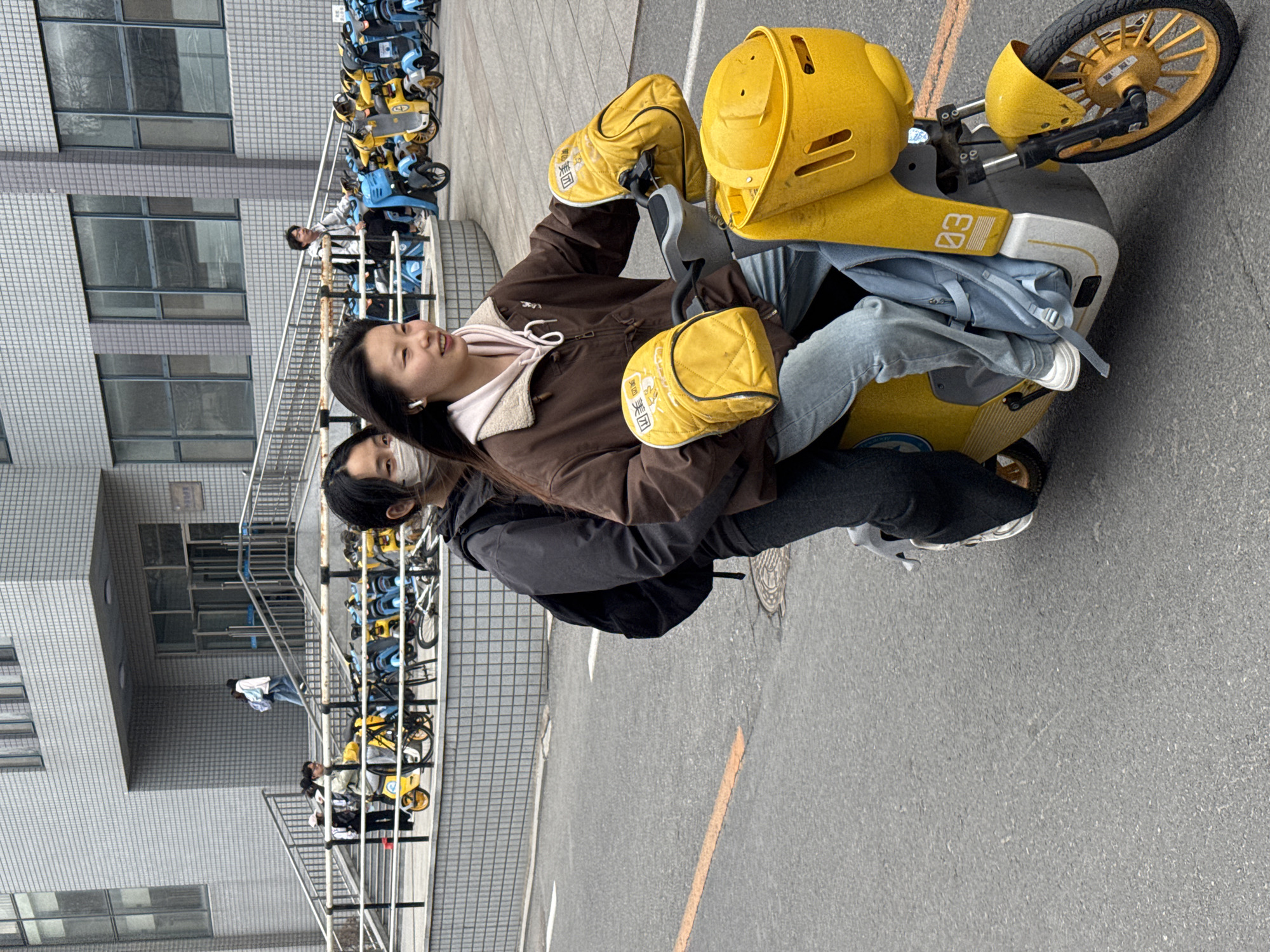 Two people are riding a yellow electric scooter on an asphalt road in Zhongtun, China. The scooter has a prominent yellow front wheel and a large yellow box-like structure in the front. The rider wears a brown jacket, jeans, and white sneakers, while the passenger wears a black jacket and gray pants. Several other scooters are parked along the left side of the road, near a gray building with multiple windows. A staircase is visible near the parked scooters.