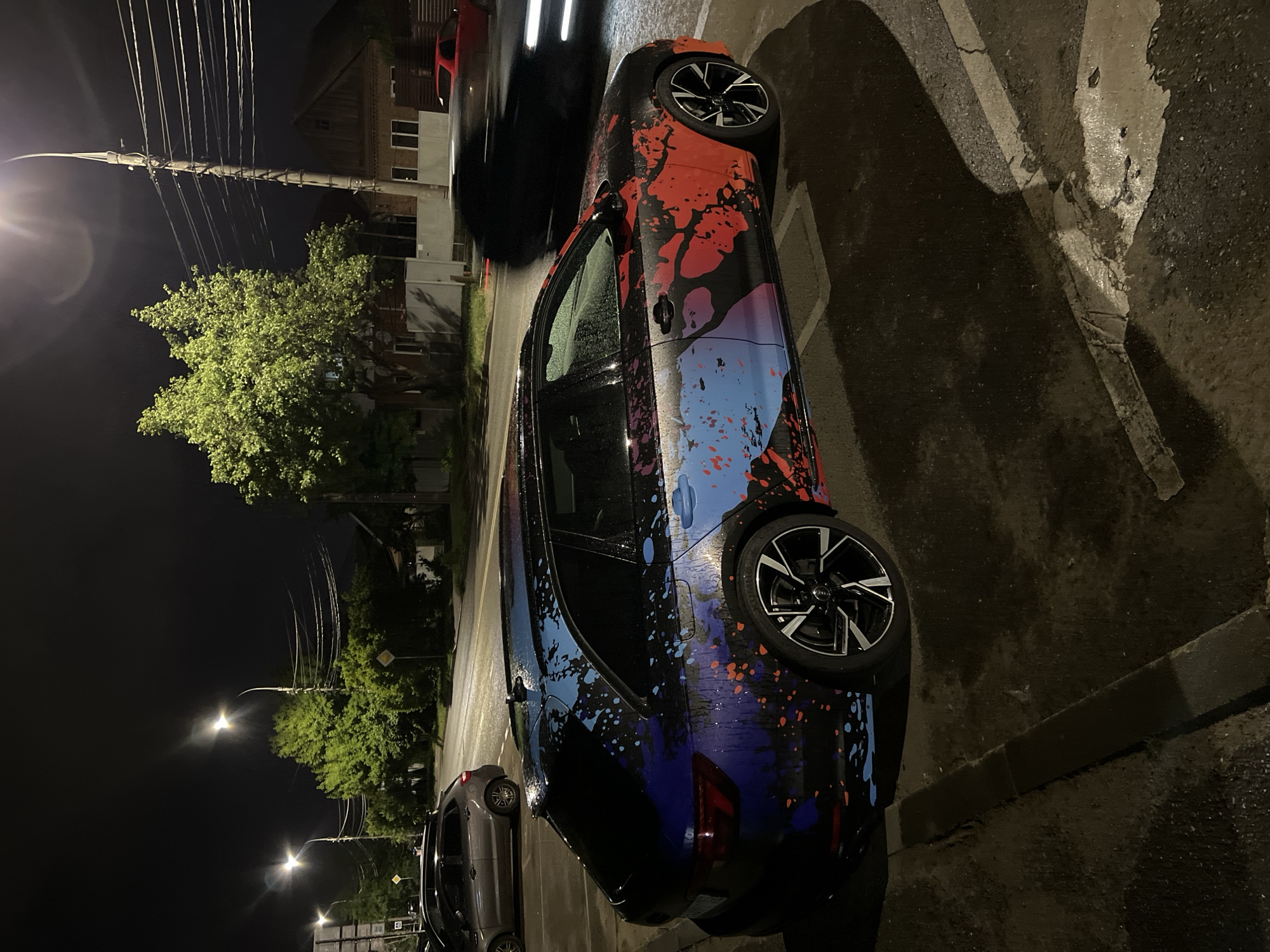This nocturnal image, taken in Krasnodar, Russia, features a strikingly customized car parked on a wet street. The vehicle, likely an Audi identifiable by its wheel cap logo, is adorned with a unique paint-splatter design transitioning from red and black at the front to blue and purple towards the rear. The tire brand "KUMHO" is visible on the rear tire.
The setting is an urban or suburban street, illuminated by several bright streetlights that cast reflections on the damp asphalt, indicating recent rain or wet conditions. White road markings and a concrete curb are visible alongside the car. In the background, lush green trees stand out under the artificial light. A prominent utility pole, laden with numerous overhead wires, rises on the left side of the frame. Dimly lit buildings, possibly residential, line the street. Another dark-colored car is parked behind the customized vehicle, and a blurry red car is visible further in the distance. No people are present in the scene. The overall impression is one of a quiet, damp night on a city street.
