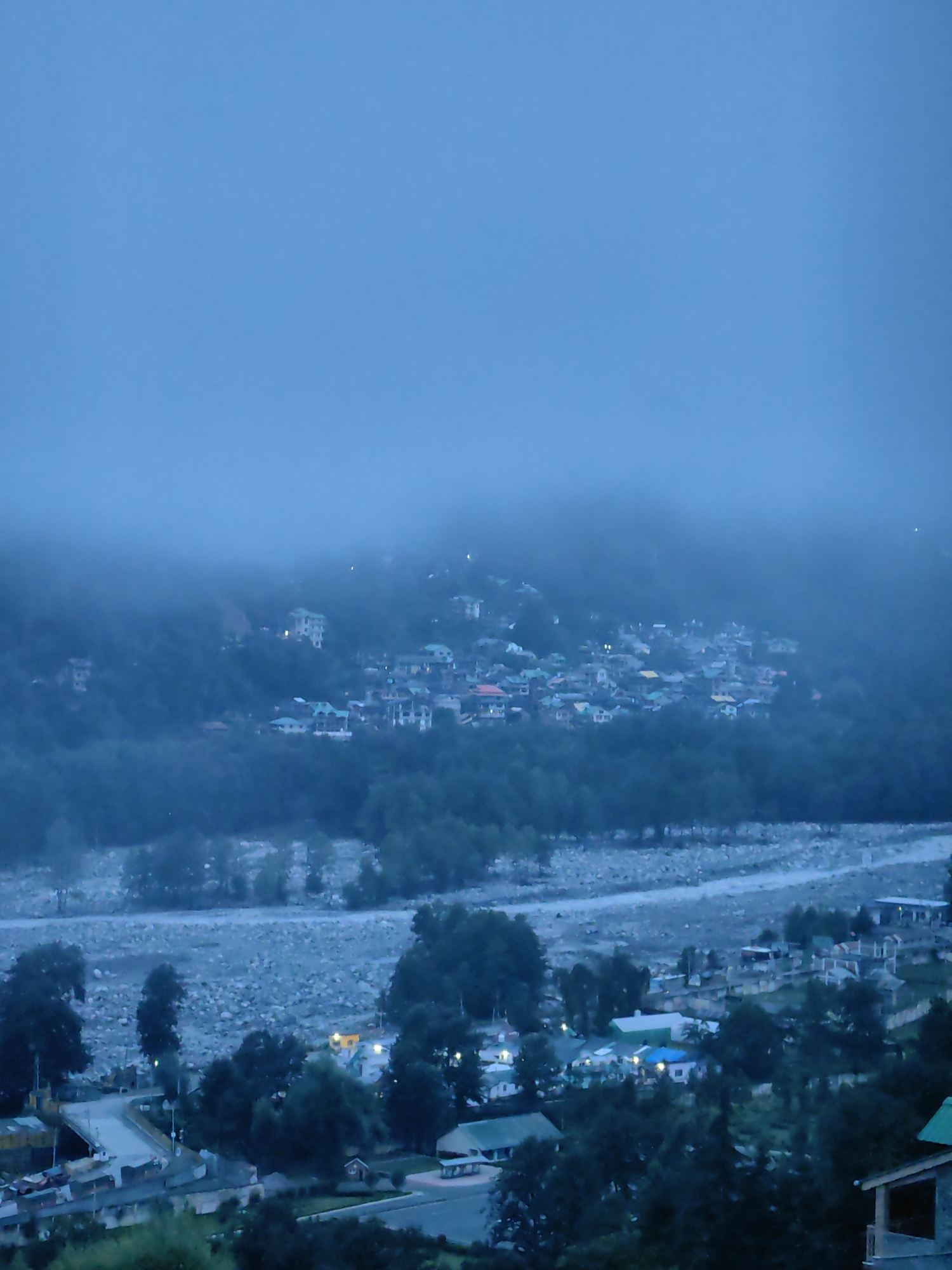 This image depicts a panoramic view of Vashisht, India, as dusk settles. The scene is dominated by a hazy, blue sky that transitions into a misty atmosphere, obscuring the distant mountains. In the midground, a densely populated village is visible, nestled on a hillside. Buildings with colorful roofs, predominantly green and yellow, are scattered amidst lush green trees. A riverbed, filled with large stones and a shallow stream, runs through the foreground, separating the village from a smaller cluster of structures. Some of these structures appear to be shops or small businesses, with awnings and lights indicating activity. A road with vehicles is also visible, suggesting a transportation network within the area. The overall impression is one of a serene, mountainous village community at the edge of day, shrouded in mist.