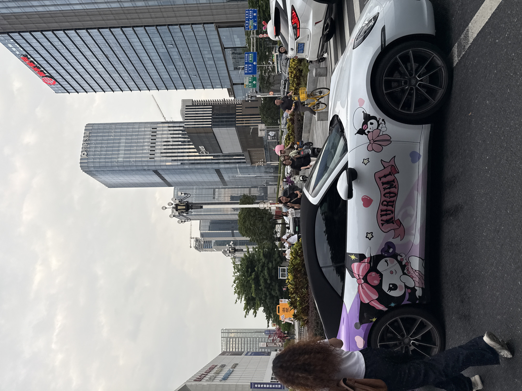 In Tianhe District, China, a white convertible Porsche is parked on a city street. The car features a custom wrap with cartoon characters, including Kuromi from Sanrio, pink bows, and hearts. A woman with curly brown hair walks away from the car, carrying a brown bag. In the background, several modern skyscrapers dominate the skyline under an overcast sky. Traffic, including cars, motorcycles, and bicycles, is visible on the street, along with pedestrians. Streetlights and traffic signs are also present.